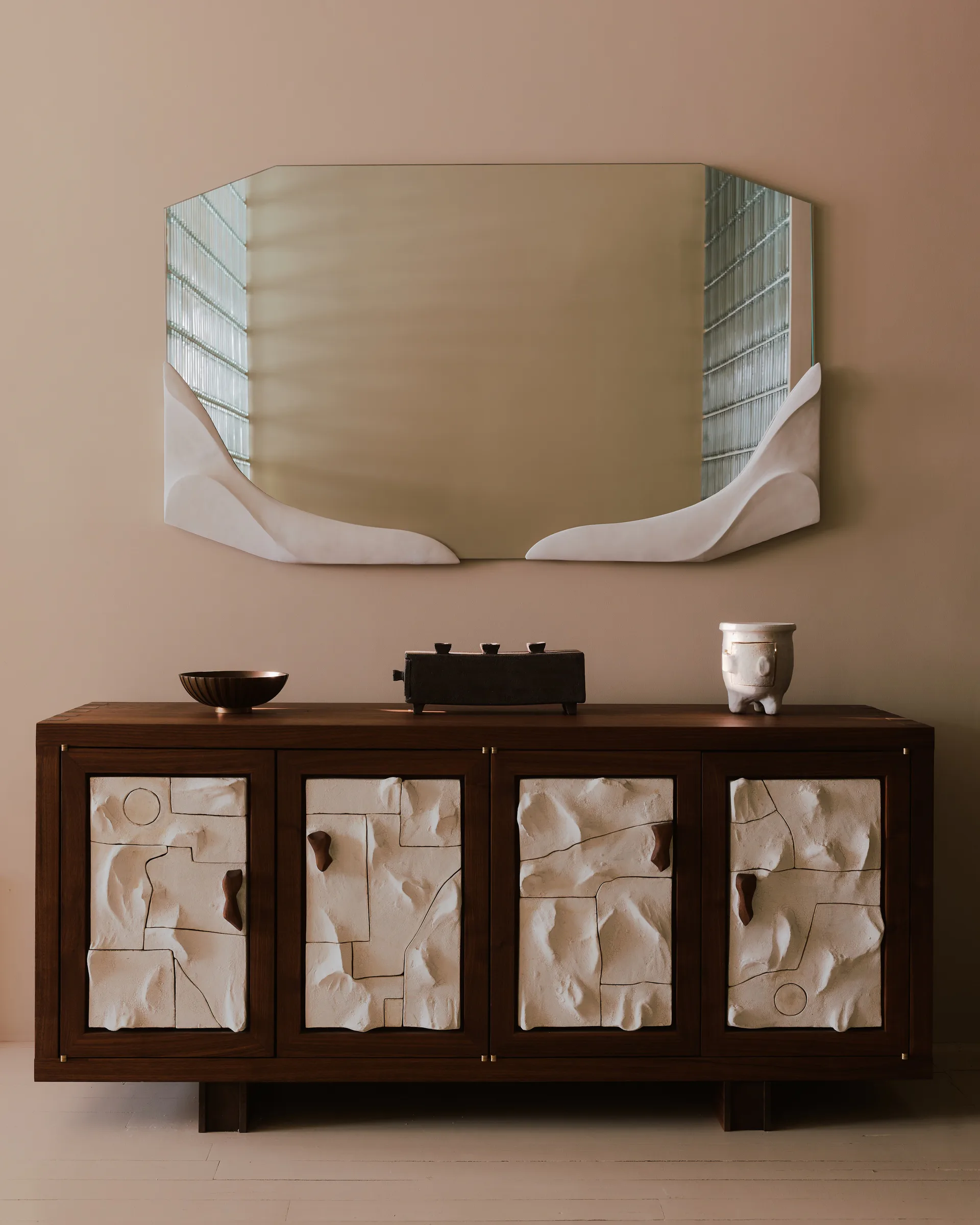 Minimalist interior featuring a dark wood credenza with four glass-front doors displaying abstract sculptural forms, topped with a black sculptural piece and ceramic vessel, beneath a large curved wall-mounted artwork, by Danny Kaplan.