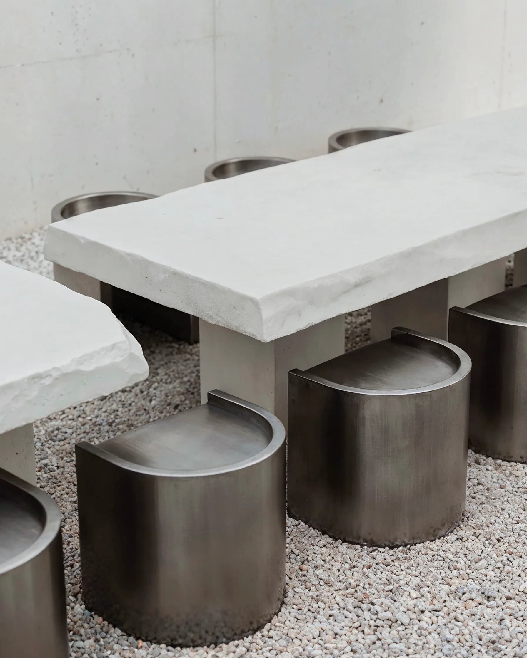 Minimalist outdoor seating area with white marble table tops and brushed metal cylindrical stools on gravel, designed by Danny Little.