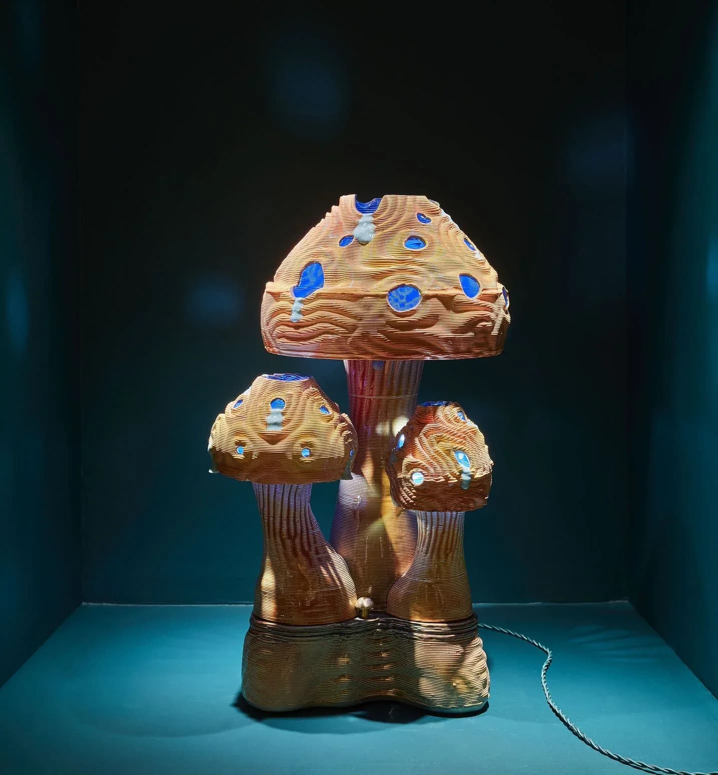 A sculptural lighting fixture featuring three stacked mushroom-shaped brass forms with blue gemstone embellishments, displayed on a turquoise surface by Dara Schaefer.