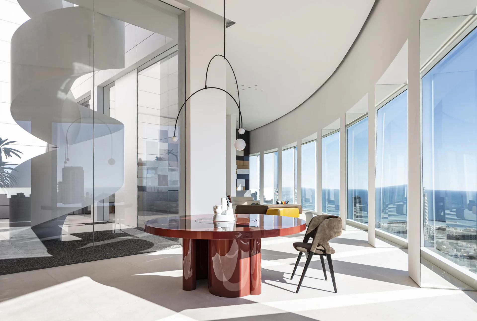 A curved high-rise dining space with floor-to-ceiling windows overlooking a cityscape, featuring a red sculptural table and modern seating by Daria Karelina.