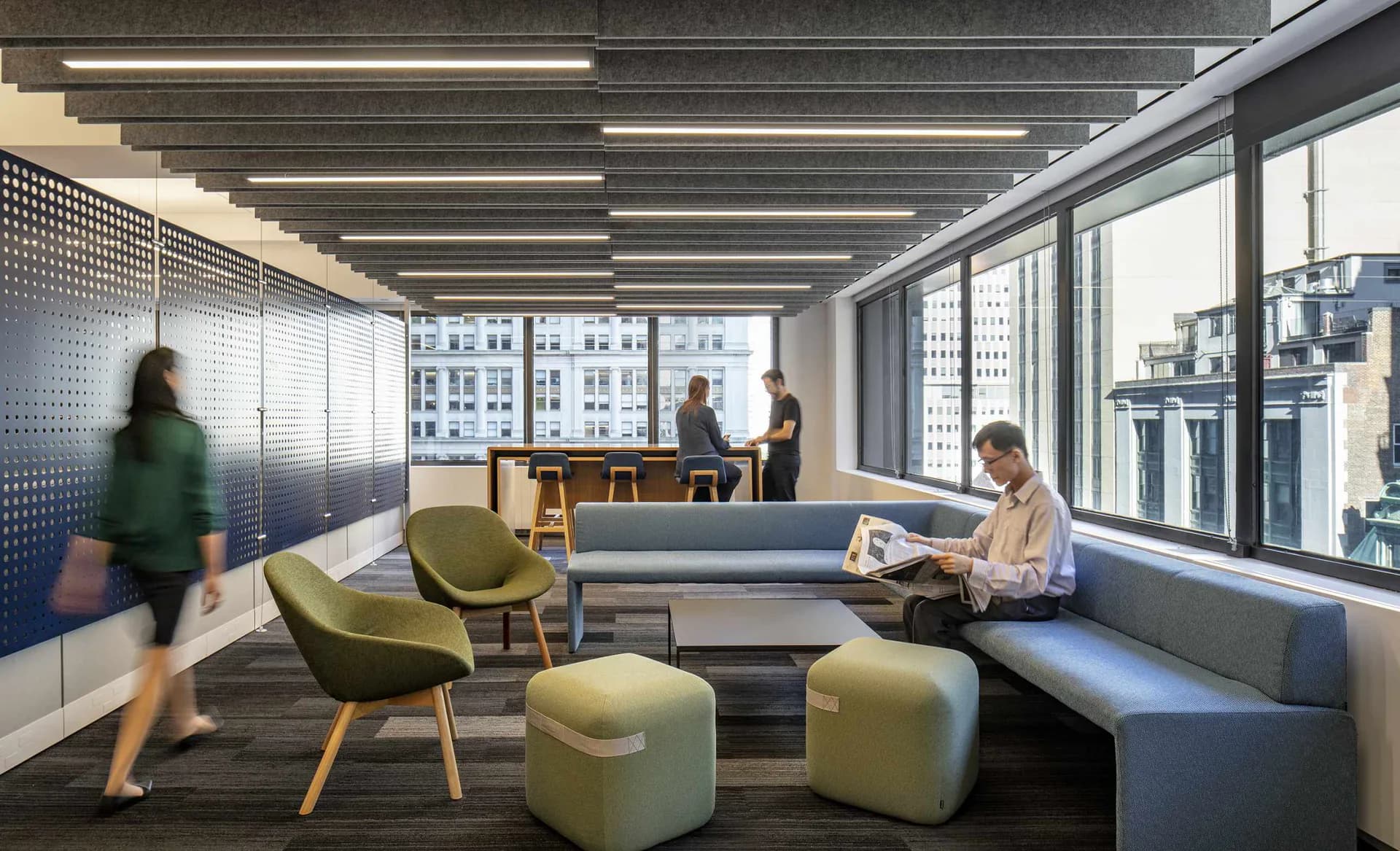 Modern office lounge with green accent chairs, blue seating, floor-to-ceiling windows overlooking city skyline, linear ceiling lights, and collaborative workspace by David Aboyoun.