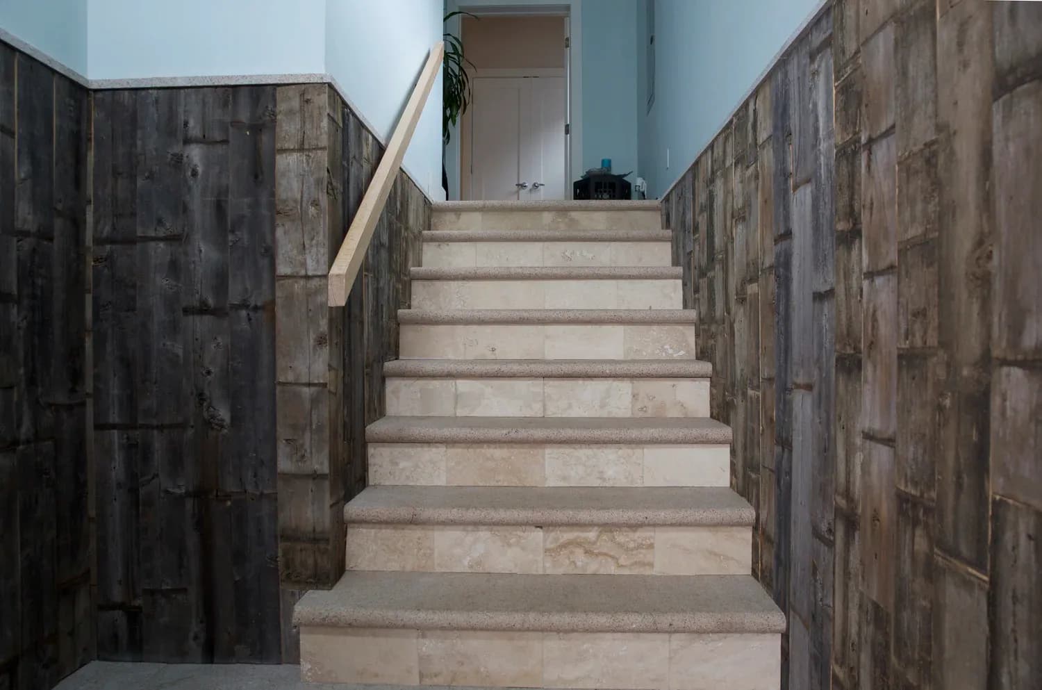 Concrete staircase with dark stone tile walls and wooden handrail leading upward to a light-filled landing by David Jones.
