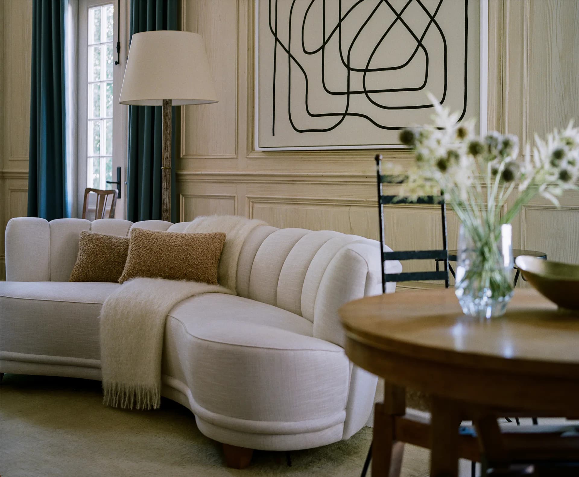 Elegant living room with curved cream upholstered sofa, warm wood side table, and abstract line art above, designed by David Lucido.