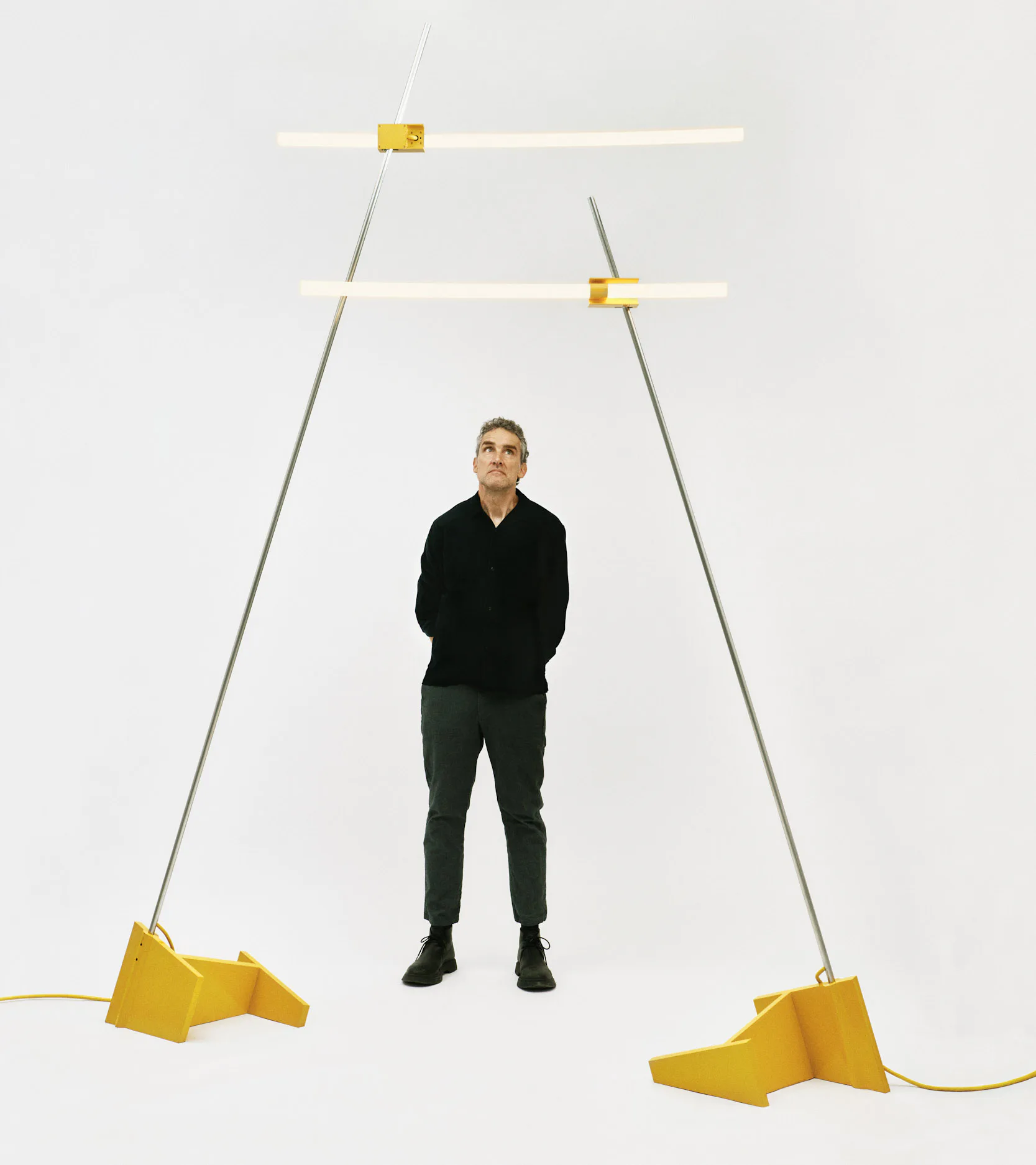Sculptural lighting installation with yellow geometric forms suspended by gray cables above a standing figure, by David Weeks.