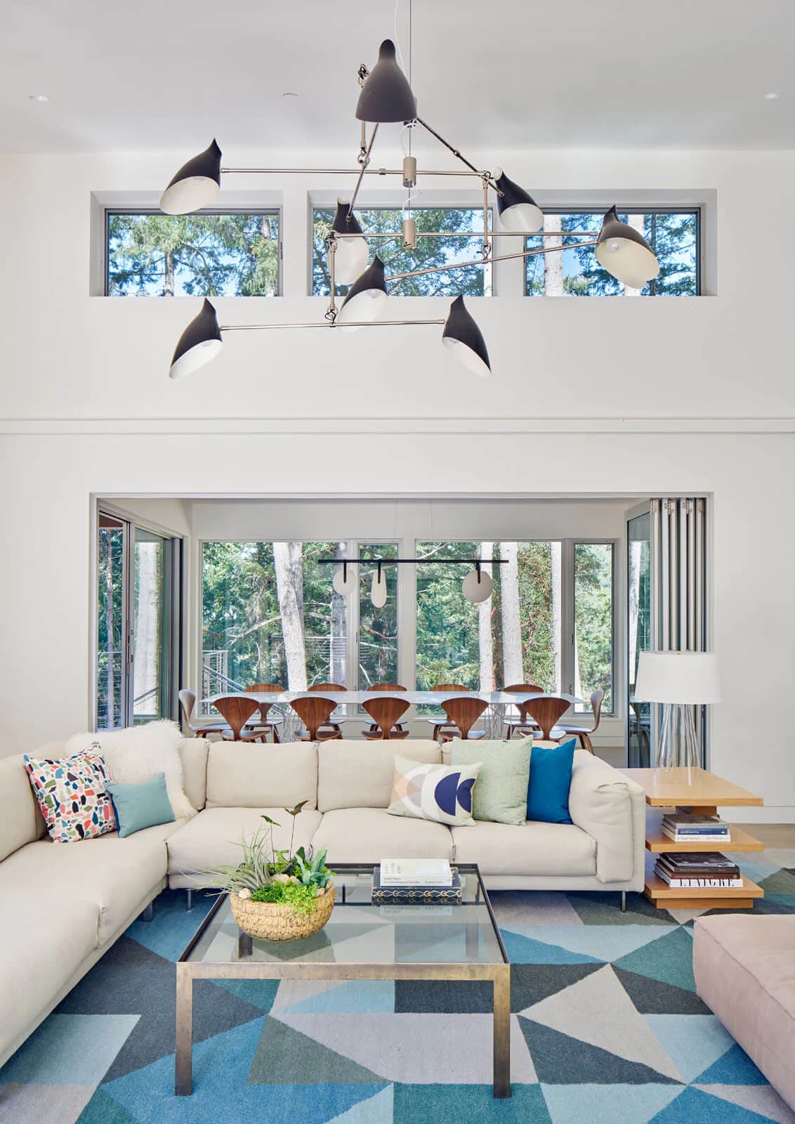 Modern living room with cream sectional sofa, geometric patterned blue area rug, glass coffee table, and sculptural black pendant chandelier by David Weeks near expansive windows overlooking trees.