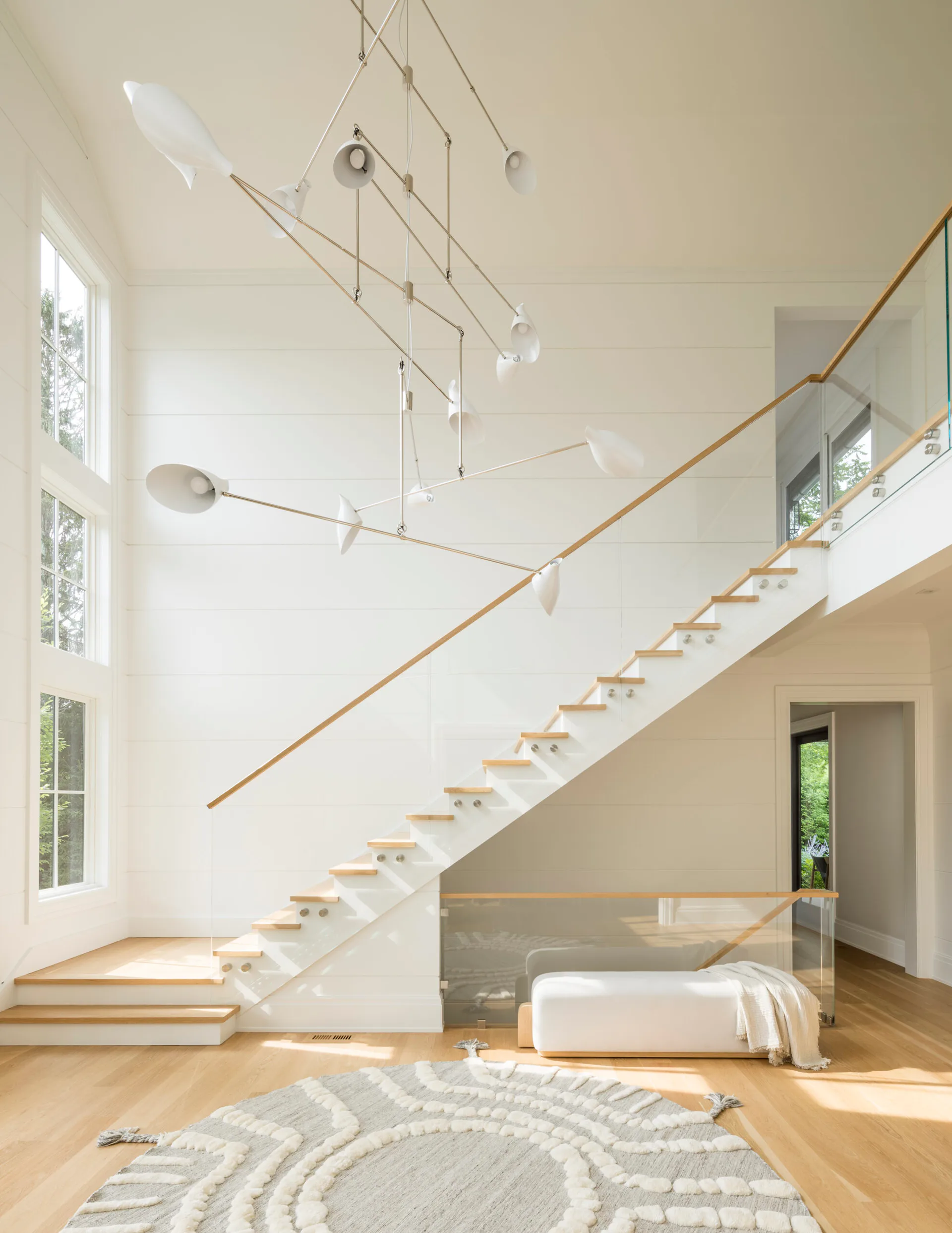 Modern double-height interior with floating wooden staircase, mobile sculptural lighting installation, warm wood floors, and bright natural light by David Weeks.