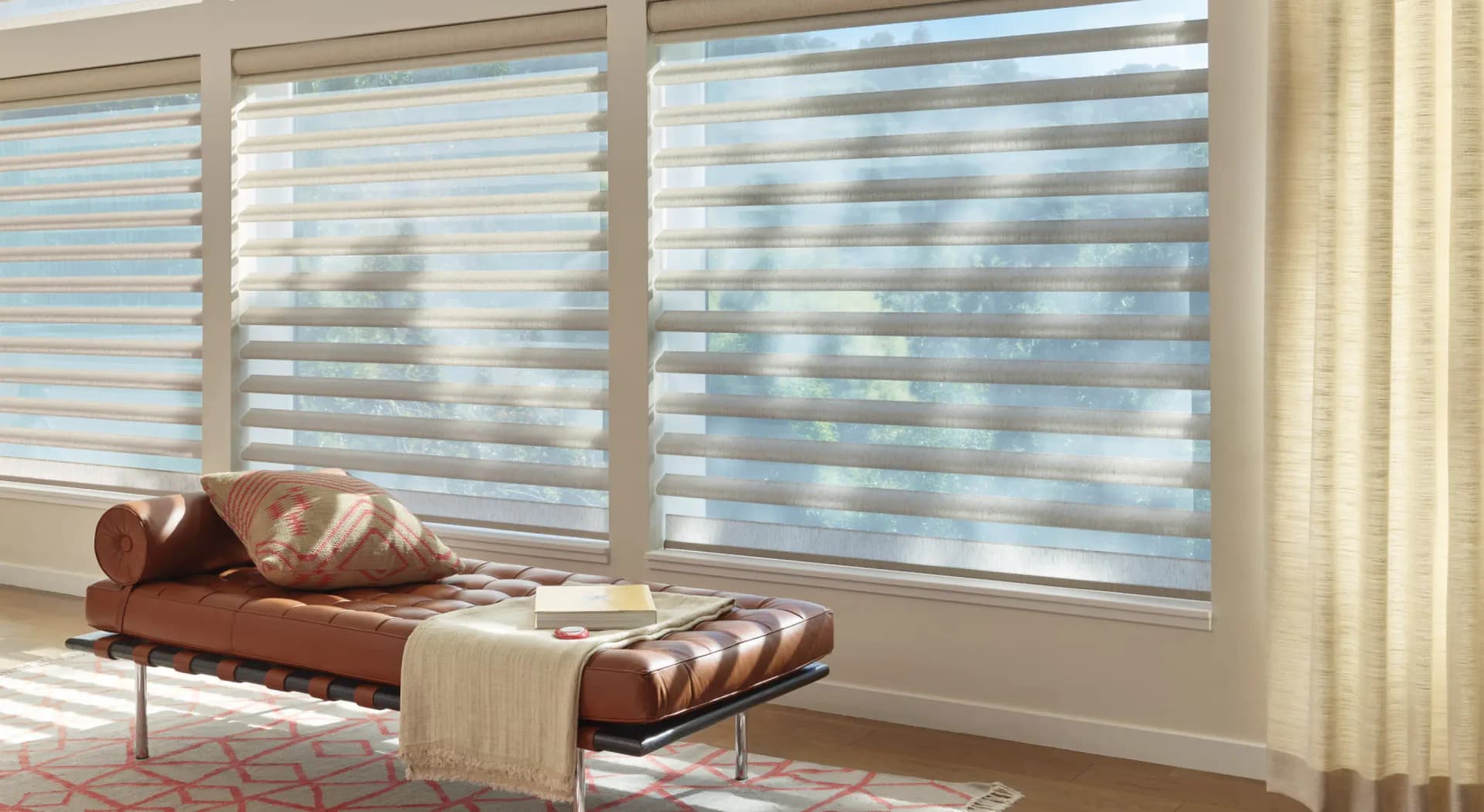 Modern living room with motorized roller shades featuring horizontal stripes, a brown leather bench with patterned cushions, and warm neutral tones by Deanna Colondona.