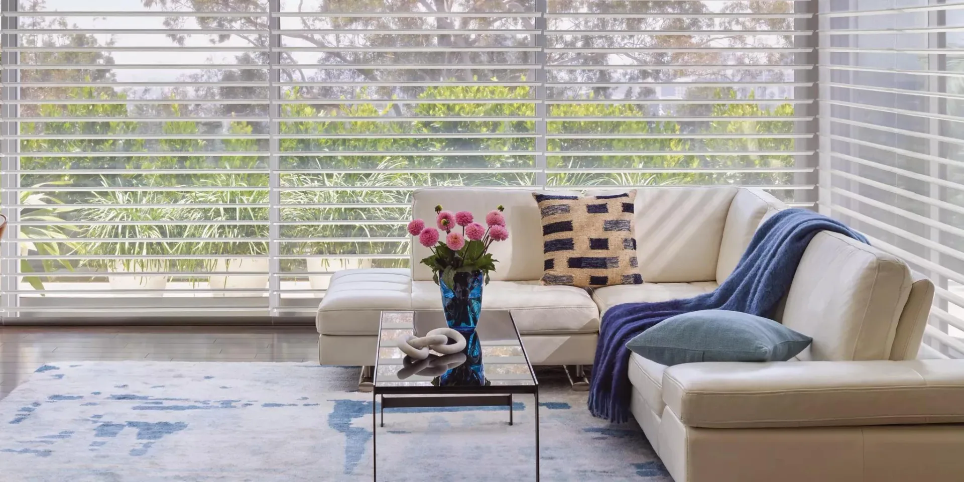 Modern living room with cream sectional sofa, blue throw blanket, glass coffee table, pink flowers, and floor-to-ceiling windows overlooking green cityscape by Deanna Colondona.