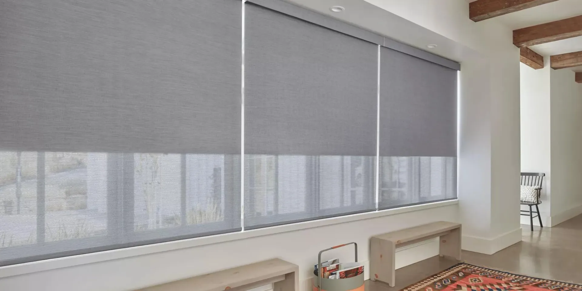 Modern living space with large windows dressed in gray roller shades, light wood benches, exposed ceiling beams, and warm neutral palette by Deanna Colondona.
