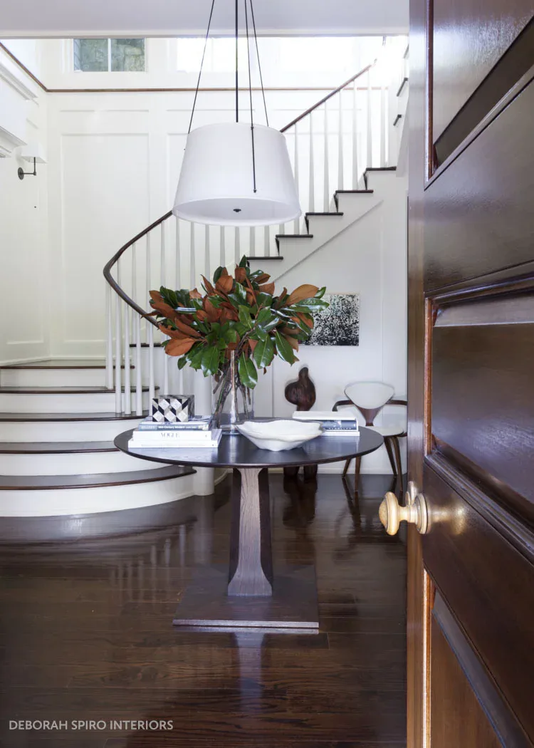 Modern foyer with curved staircase, dark wood flooring, white railings, round pedestal table with orange floral arrangement, and pendant lighting by Debbie Spiro.