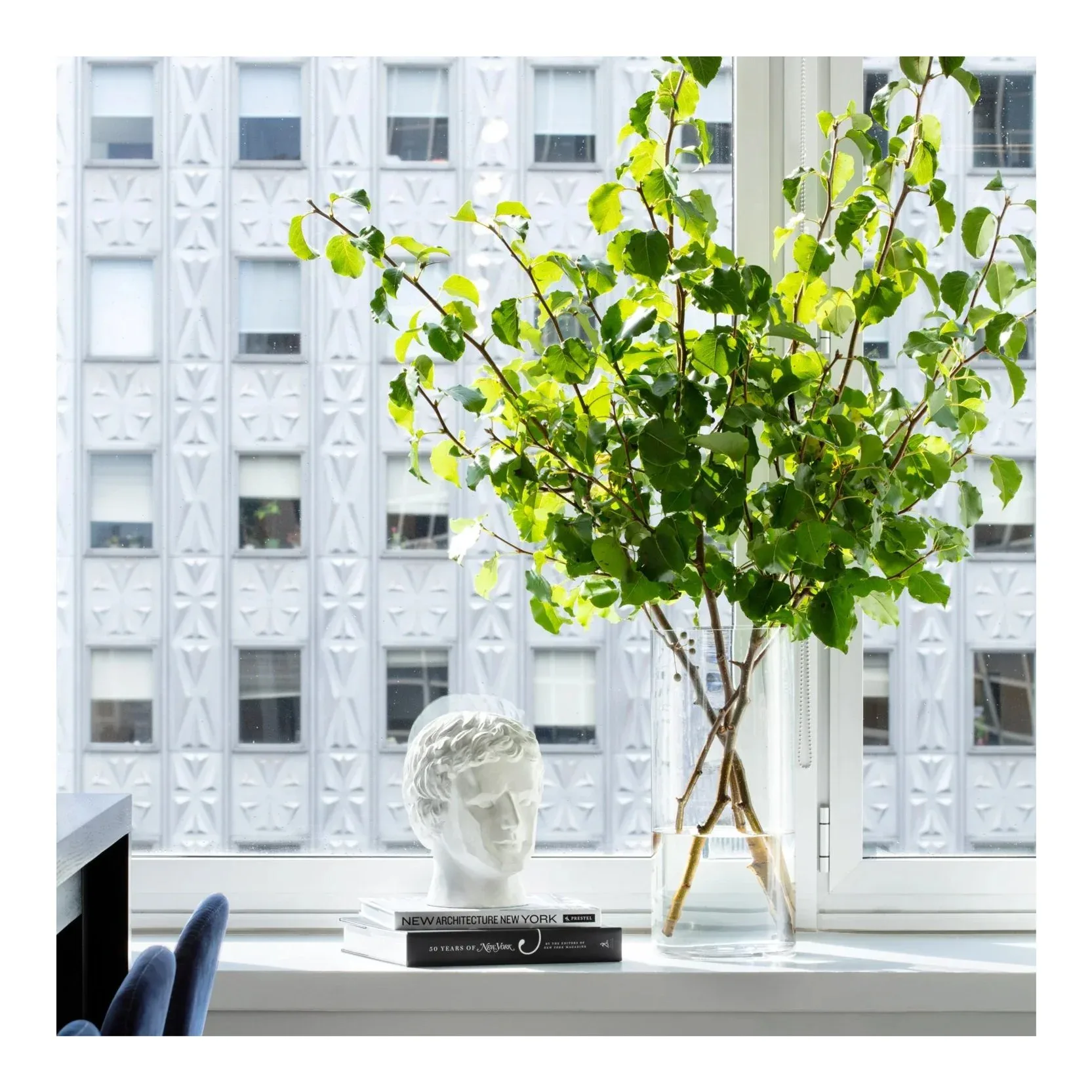Bright minimalist interior with white ornamental wall paneling, fresh green foliage arrangement in glass vase, white classical bust sculpture, and blue accent chair by Debbie Spiro.
