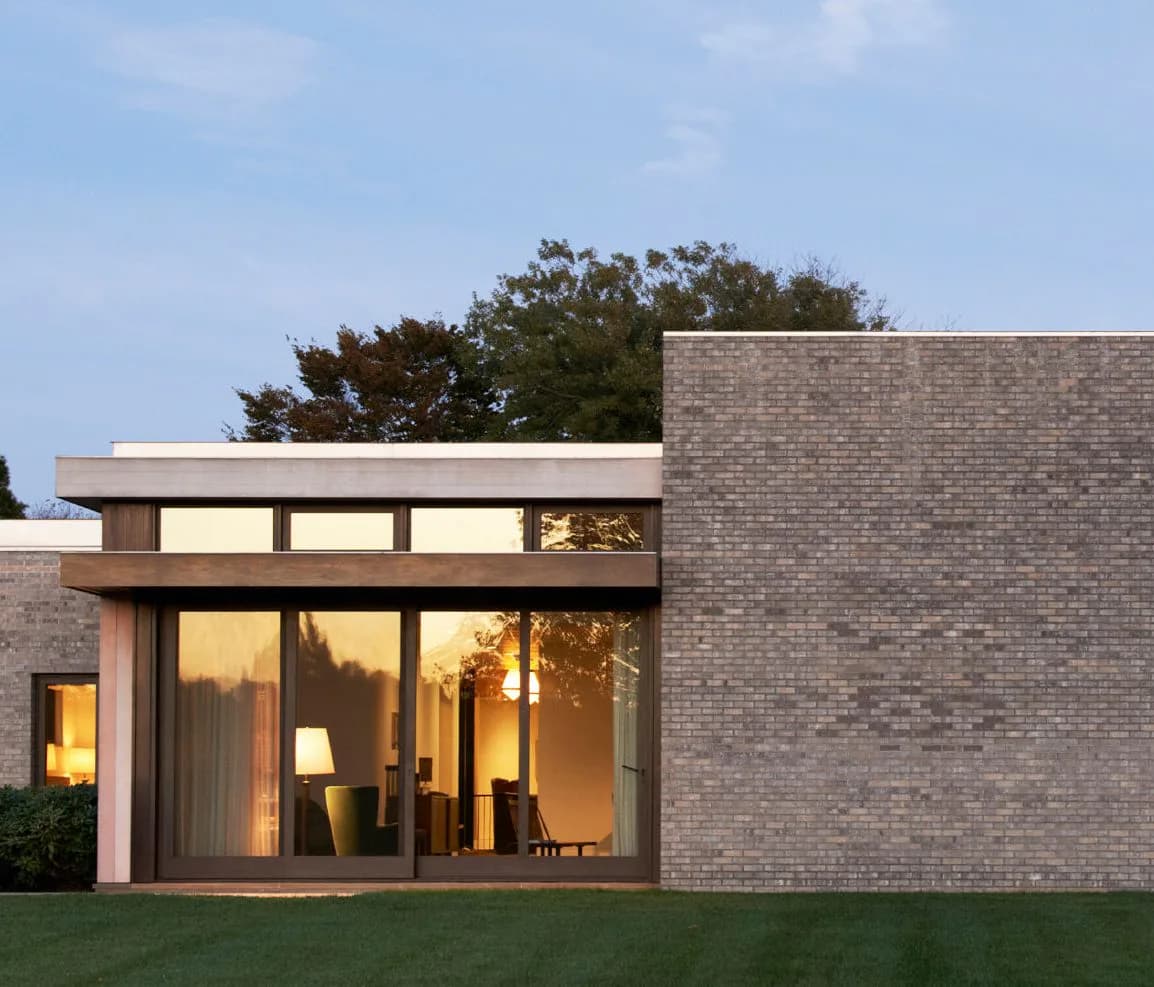 Modern brick residential building with large glazed corner windows, illuminated interior, and manicured lawn at dusk by Deborah Berke.