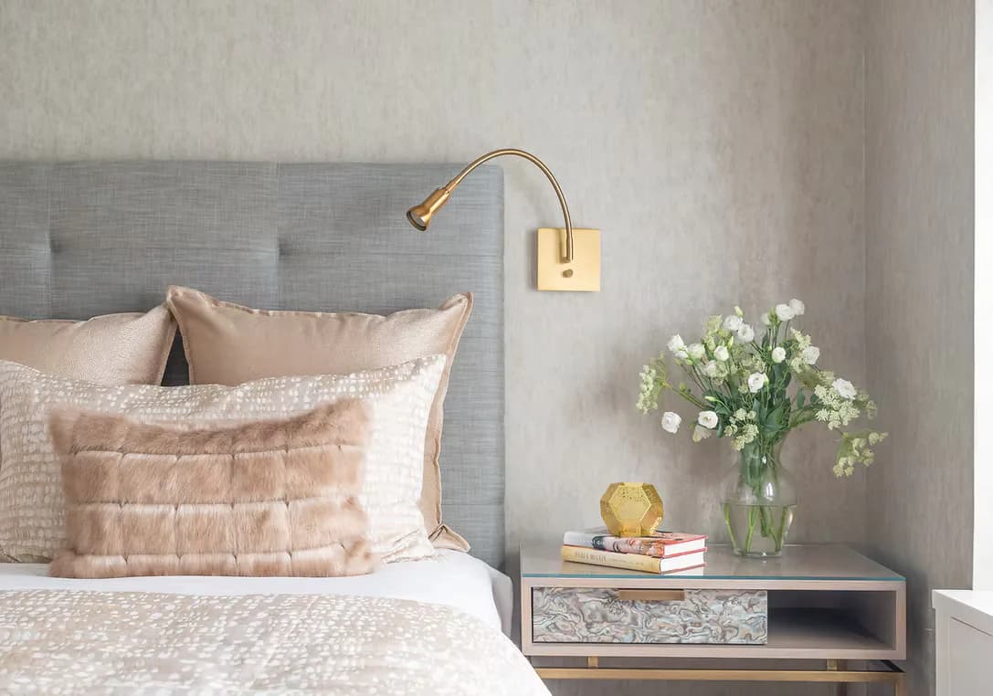 Modern bedroom interior featuring a gray upholstered headboard, blush velvet pillows, brass swing-arm wall sconce, and a wooden nightstand with floral arrangement by Deborah Emery.
