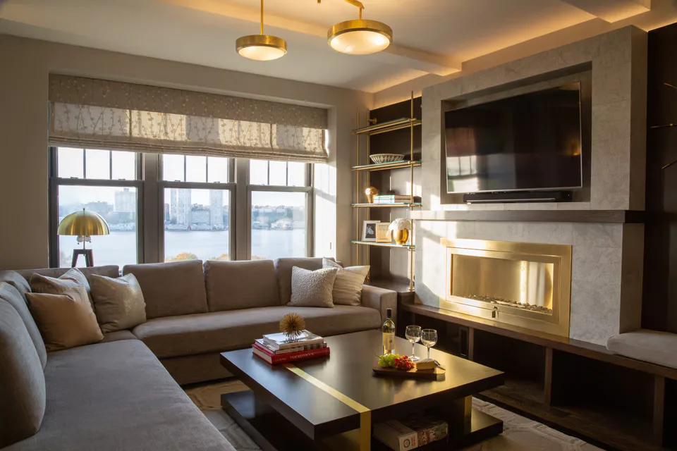 Modern living room with taupe sectional sofa, linear fireplace with brass surround, floor-to-ceiling windows overlooking water, and integrated shelving by Deborah Emery.