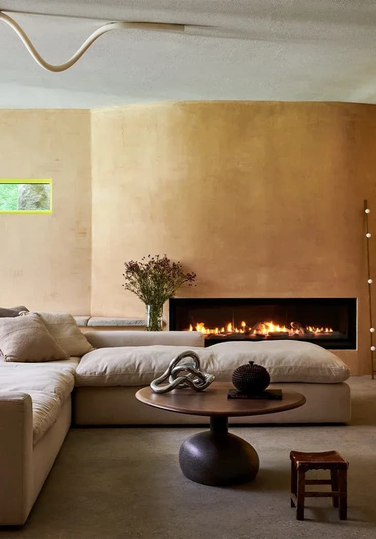 Minimalist bedroom with warm terracotta walls, linear fireplace with glowing flames, low platform bed with pillows, sculptural round side table, and sparse furnishings by Delia Kenza.