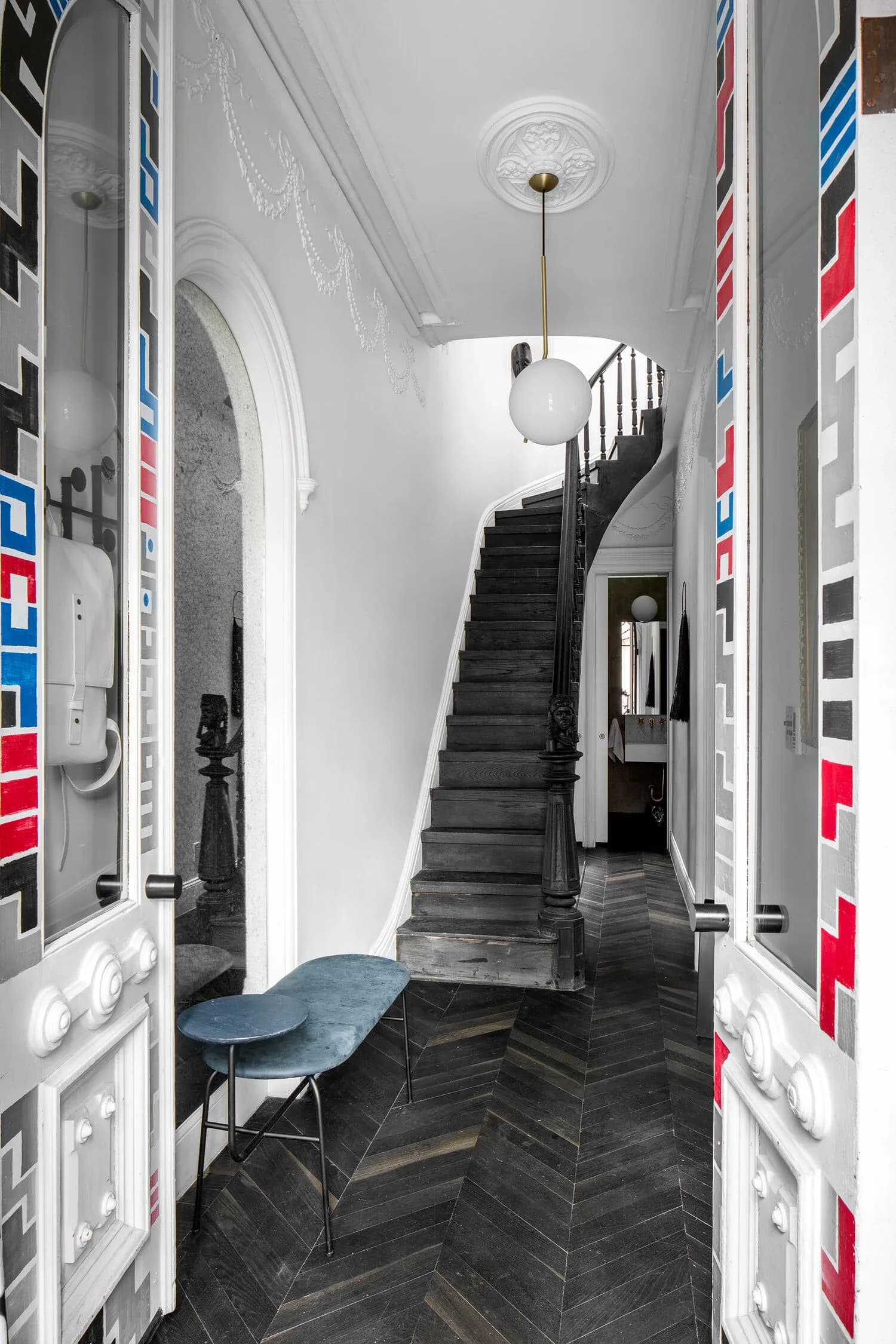 Modern stairwell with dark herringbone flooring, white walls, and colorful striped wall panels, featuring a pendant light and blue chair by Delia Kenza.