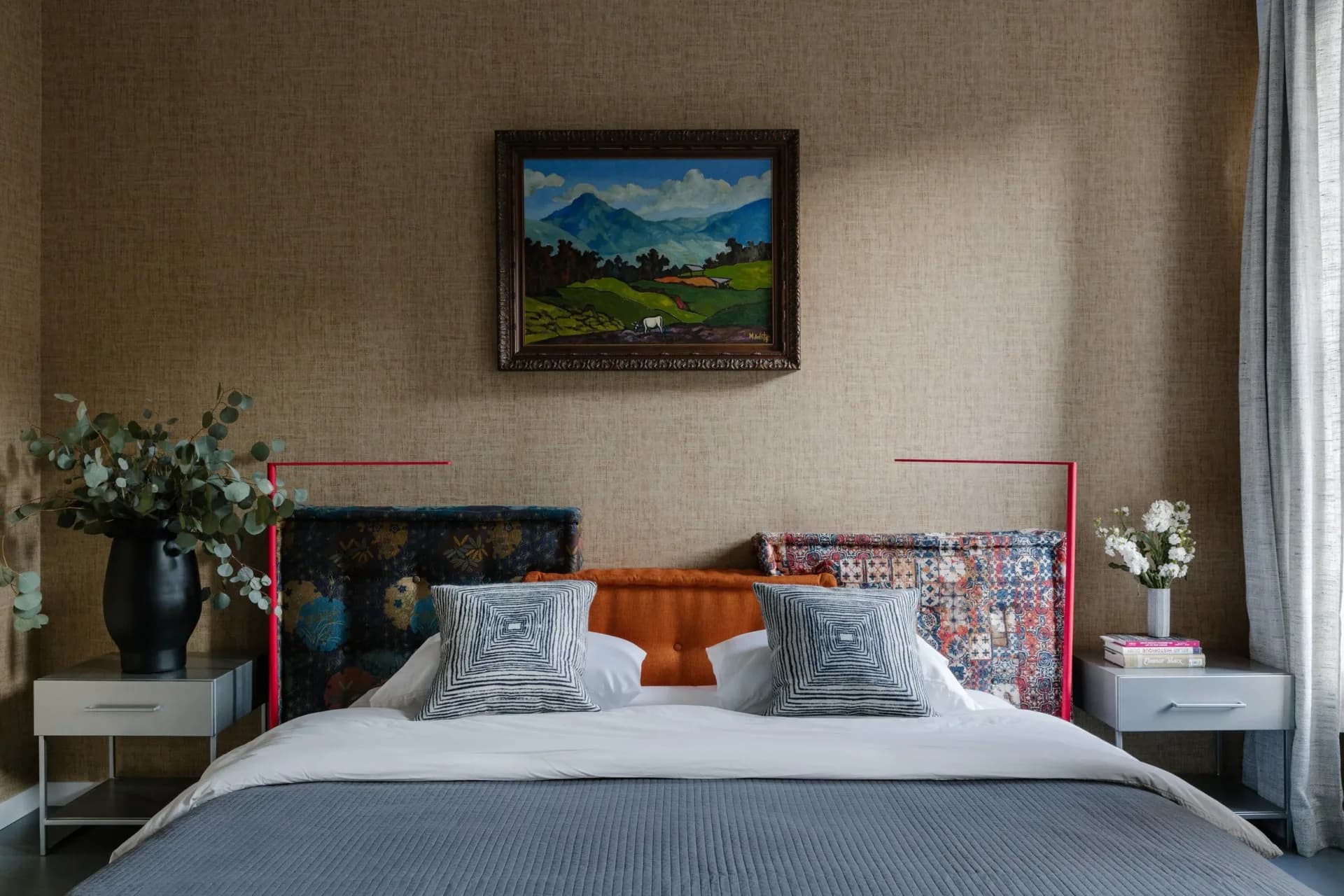 Modern bedroom with colorful upholstered headboard, layered bedding, geometric pillows, neutral walls, landscape artwork, and coordinating nightstands by Delia Kenza.