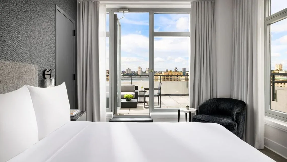 Modern hotel bedroom with white bedding, grey upholstered headboard, and floor-to-ceiling windows overlooking a cityscape by Derrick Sibley.