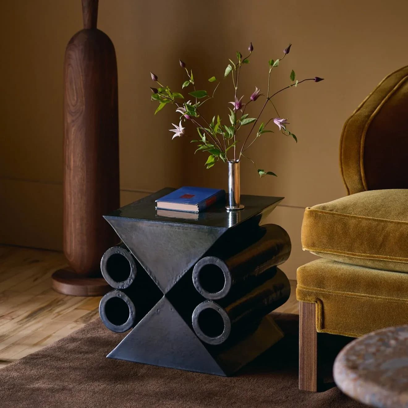 Contemporary interior with geometric black side table featuring cylindrical legs, styled with floral arrangement, blue books, and mustard velvet seating by Devin Wilde.