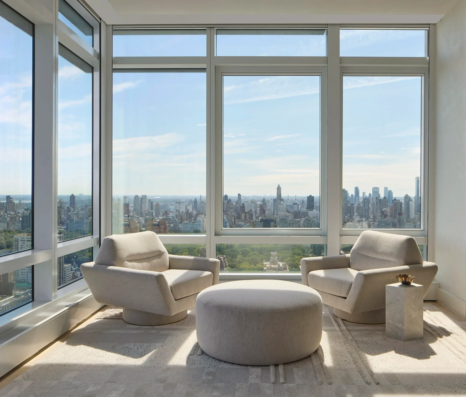 Modern high-rise penthouse with floor-to-ceiling windows overlooking Manhattan skyline, featuring cream upholstered lounge chairs and rounded ottoman in minimalist composition by Diana Litchfield.