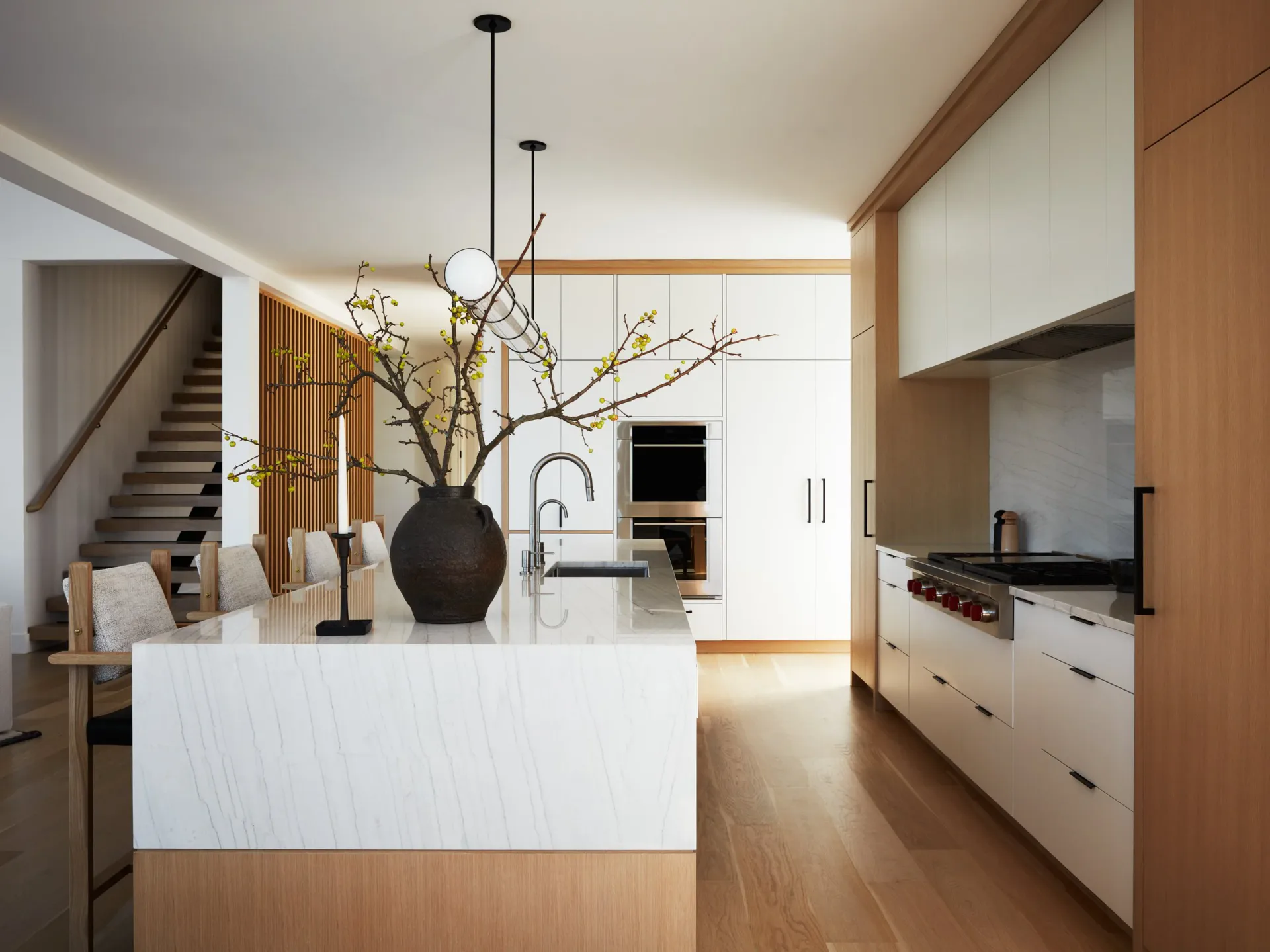 Modern kitchen and dining space with white cabinetry, warm wood accents, and a sculptural black vase with branches on the island, designed by Diana Litchfield.
