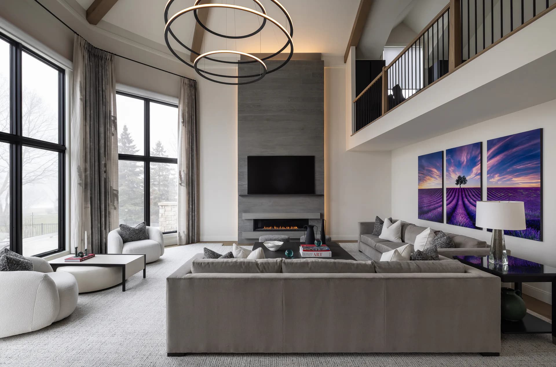 Modern two-story living room with gray sectional sofa, floor-to-ceiling windows, floating mezzanine, and purple landscape artwork by Diana Reinlein.