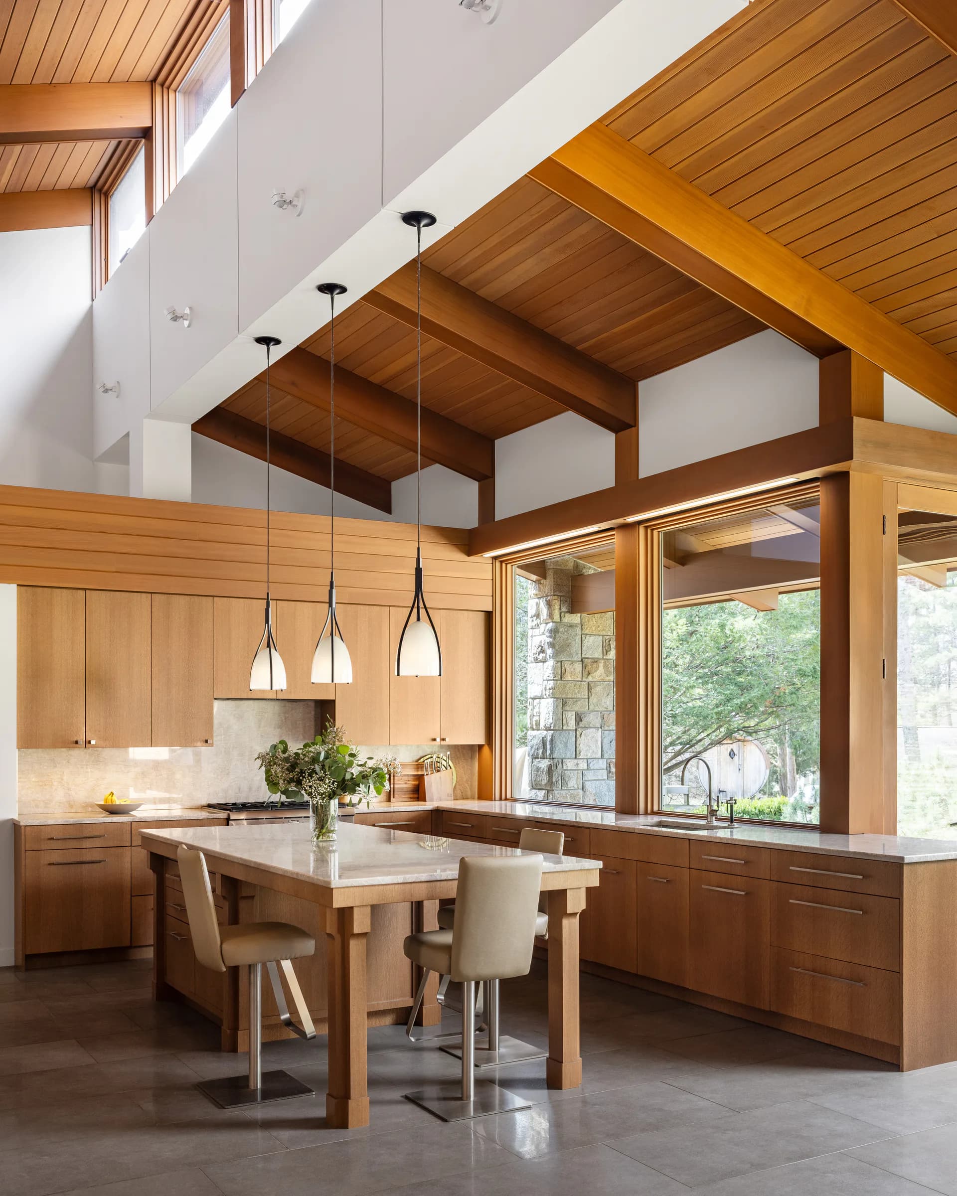 Modern kitchen with warm wood cabinetry, white countertops, and pendant lighting beneath exposed wooden ceiling beams overlooking large windows by Diana Reinlein.