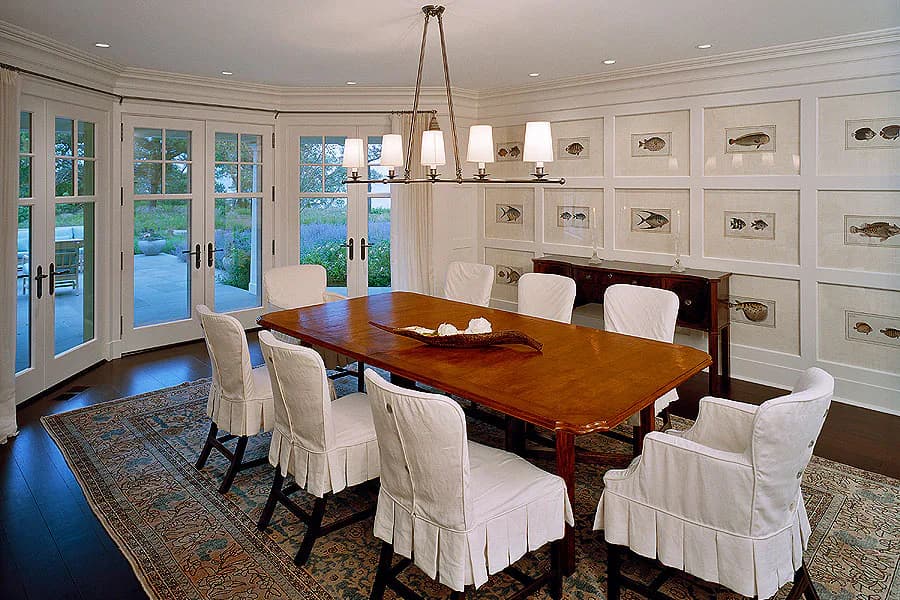 Elegant dining room with mahogany table, cream slipcovered chairs, coffered walls with hardware displays, large glass doors, and pendant lighting by Diana Sawicki.