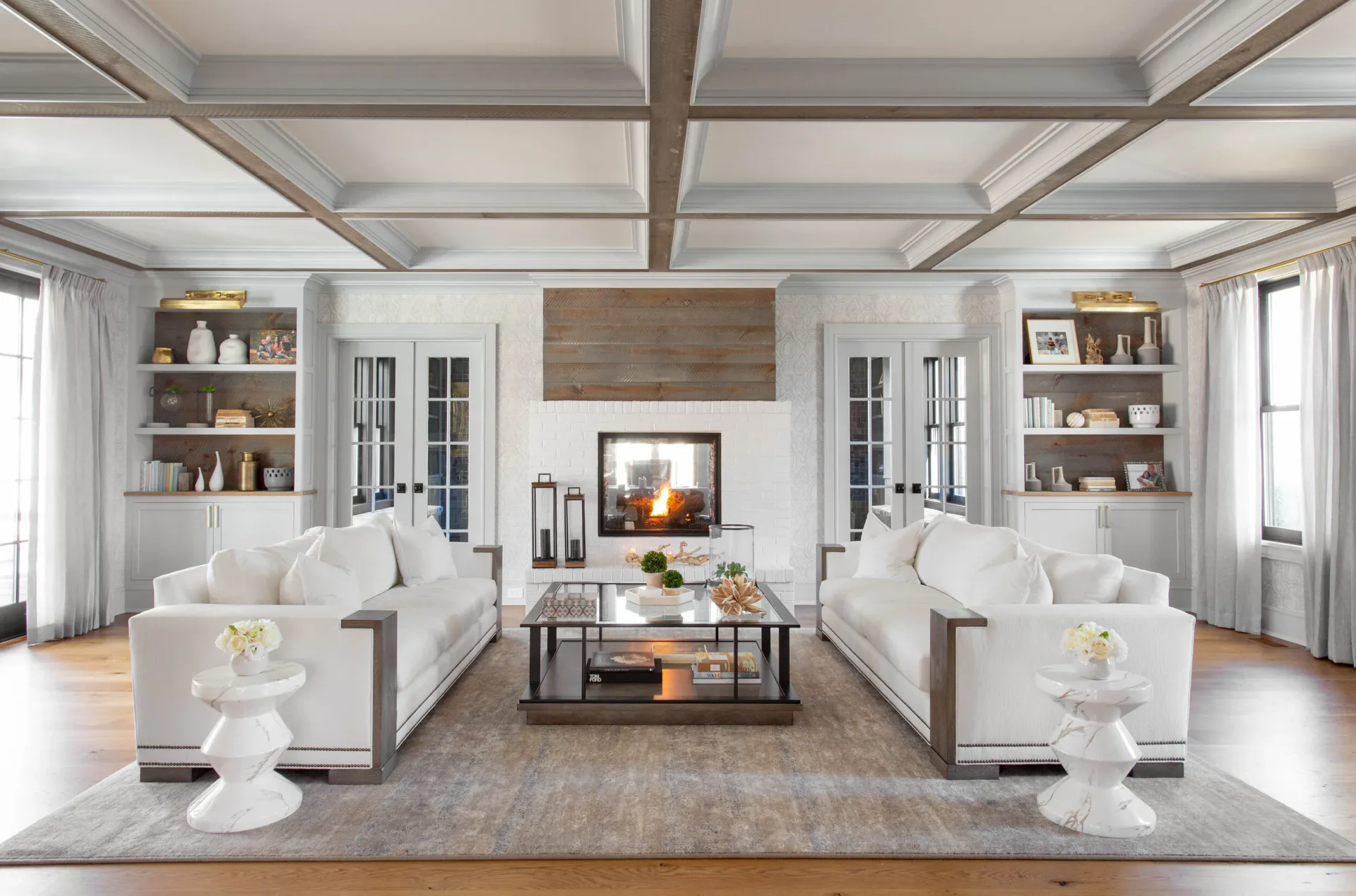 Symmetrical modern living room with white sofas, coffered ceiling, built-in shelving, central fireplace, and polished wood floors by Diane Guariglia.