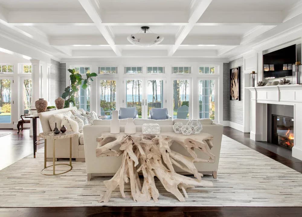 Bright modern living room with cream sectional sofa, driftwood coffee table, coffered ceiling, fireplace, and walls of glass doors overlooking water by Diane Guariglia.