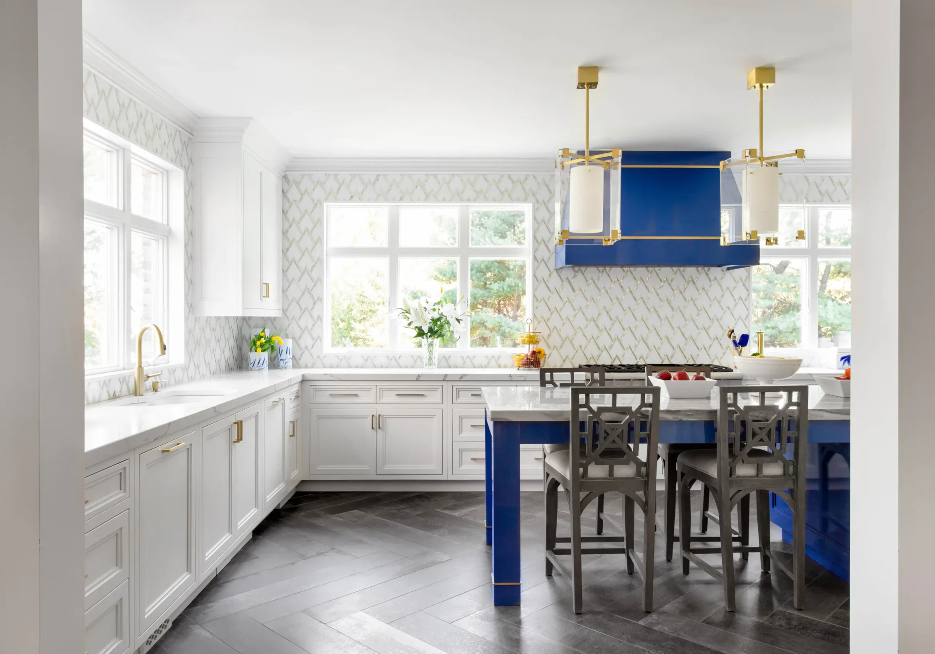Modern kitchen with white cabinetry, marble herringbone backsplash, and blue island seating area with brass pendants by Diane Guariglia.