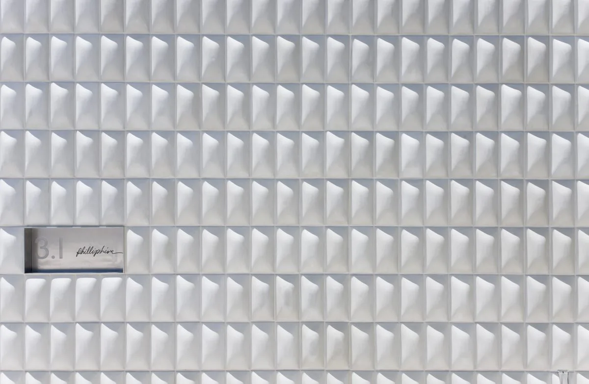 Minimalist interior wall featuring repeated geometric white panel elements with subtle shadows, creating a textured surface design by Dina Reziapova.
