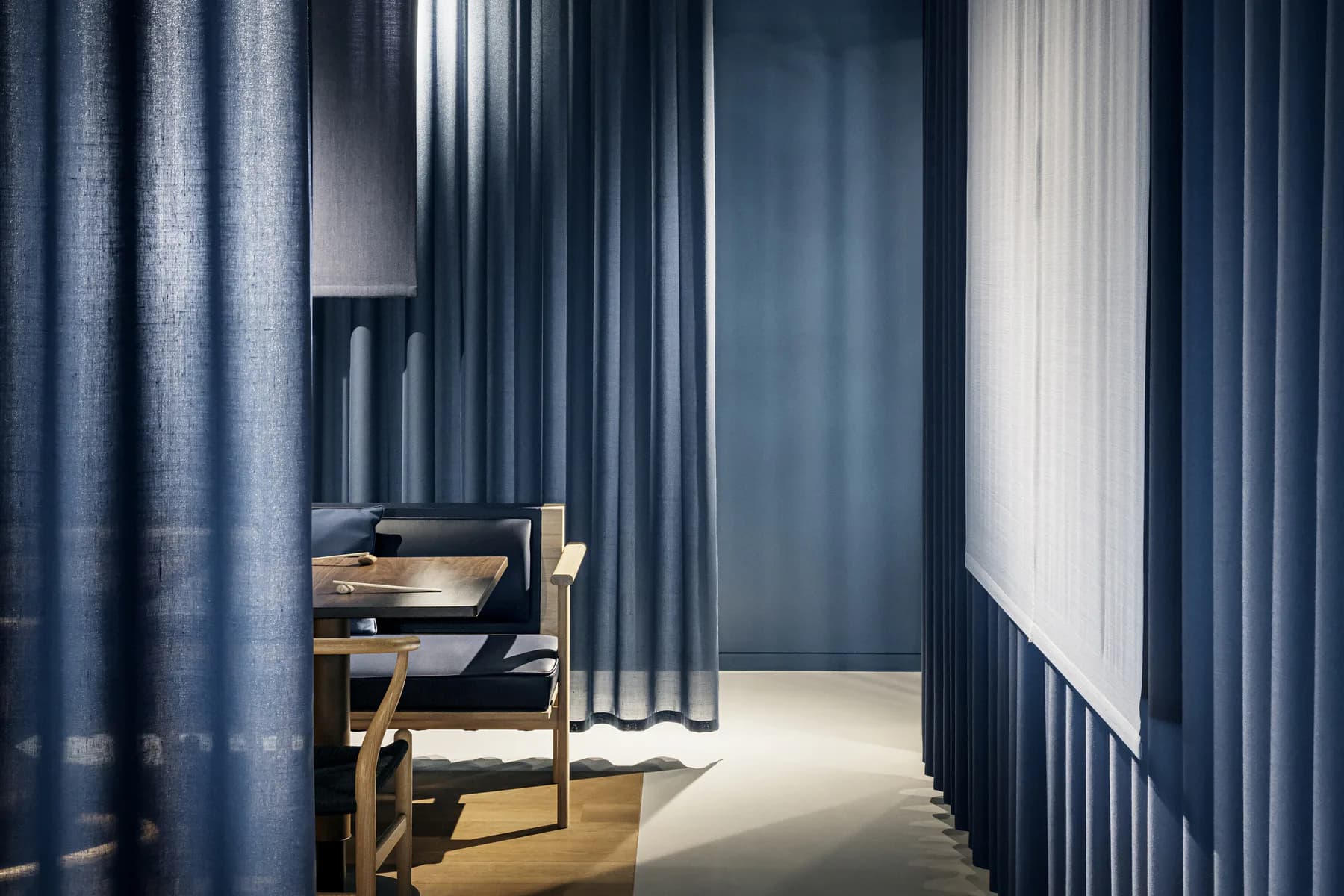 A modern interior space featuring floor-to-ceiling navy blue curtains, wooden furniture, and natural light from windows, creating a minimalist design by Ditte Hesse.