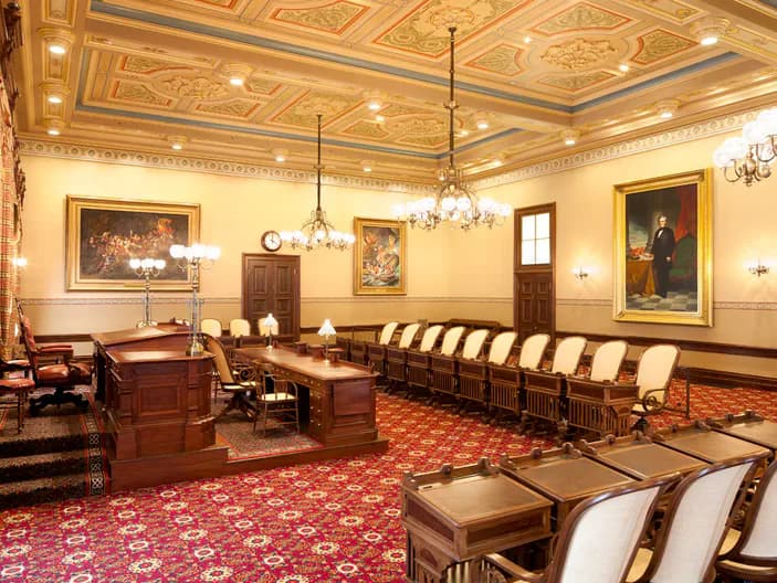 Elegant neoclassical meeting room with coffered ceiling, burgundy patterned carpet, wood paneling, and formal seating arrangement designed by Domingo González.