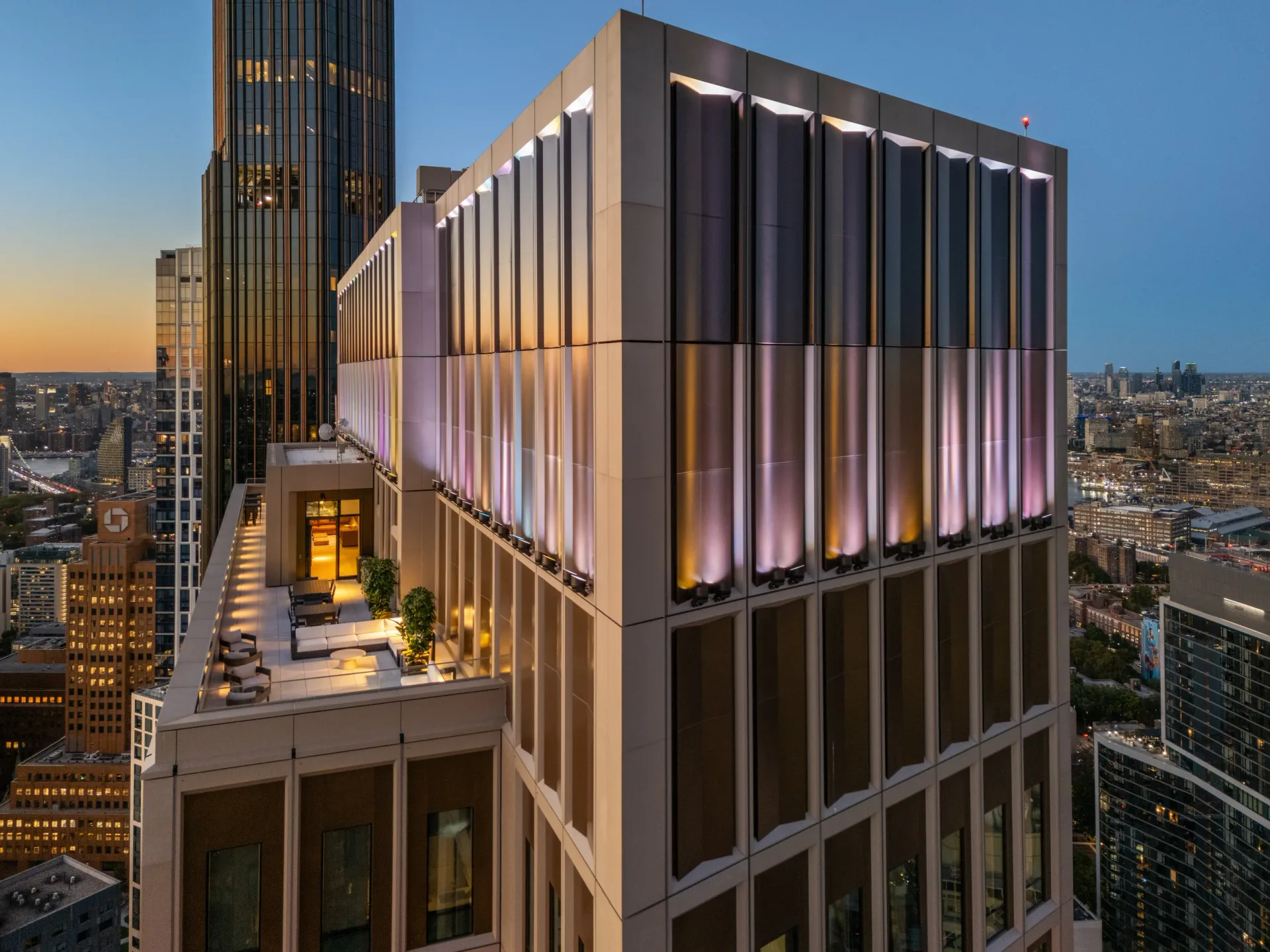 Contemporary high-rise building facade at dusk with illuminated vertical bronze louvers and glass panels overlooking a cityscape, designed by Dominic Kozerski.