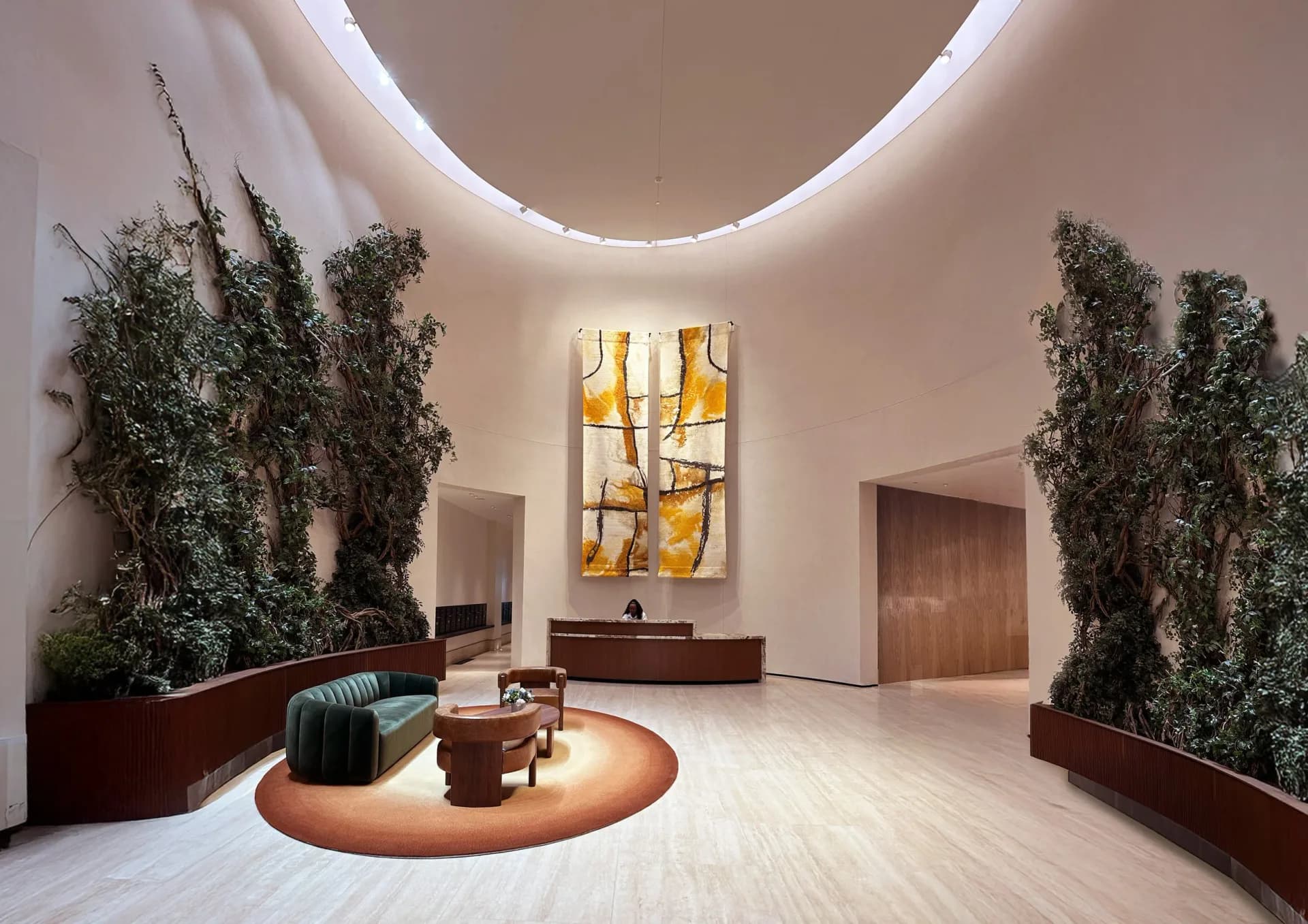 Modern circular gallery space with curved ceiling light, tall potted plants, teal armchair, wooden side table, and abstract golden wall art by Dominic Kozerski.