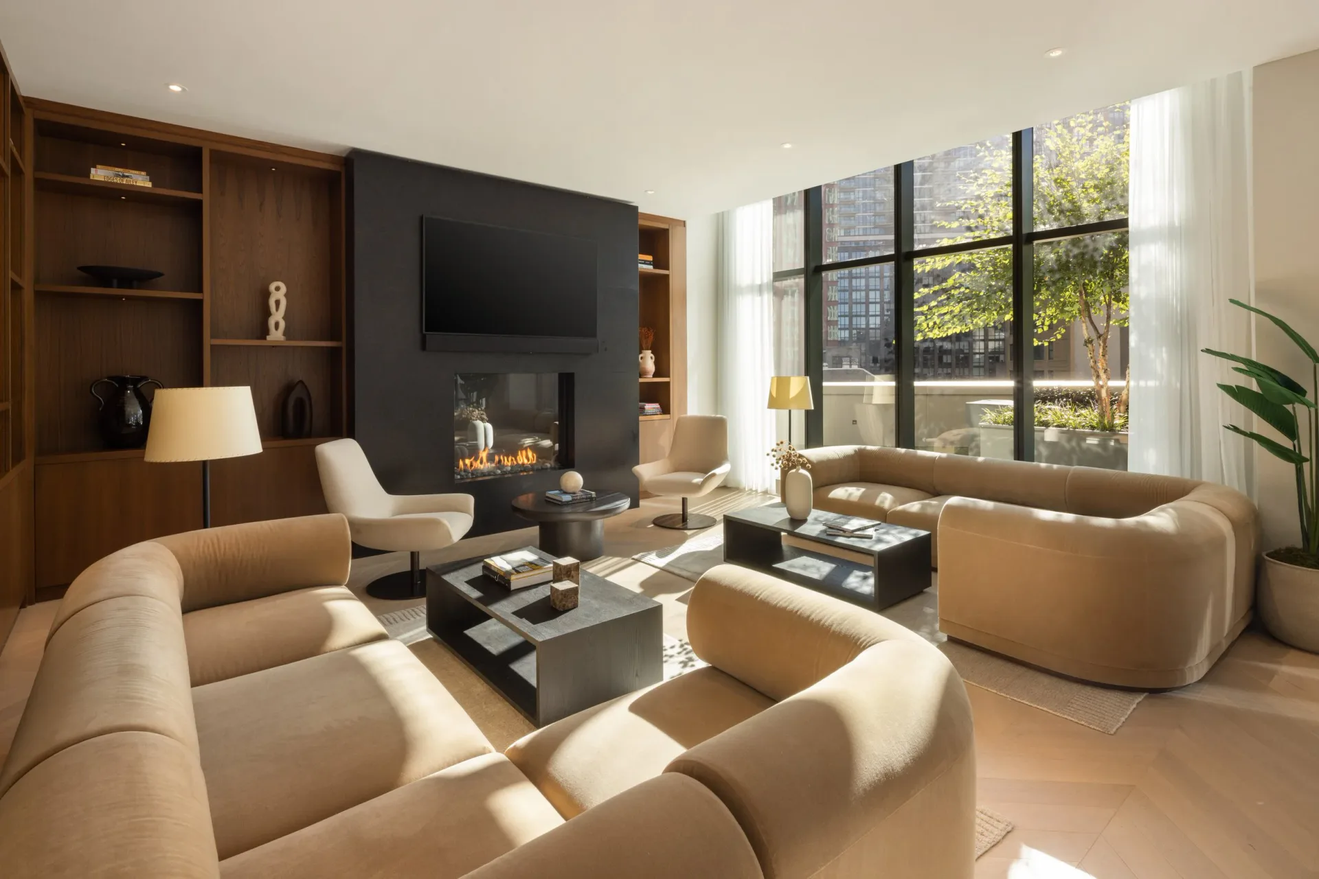 Modern living room with curved tan seating, dark accent wall with fireplace, wood shelving, and floor-to-ceiling windows overlooking greenery by Dominic Kozerski.