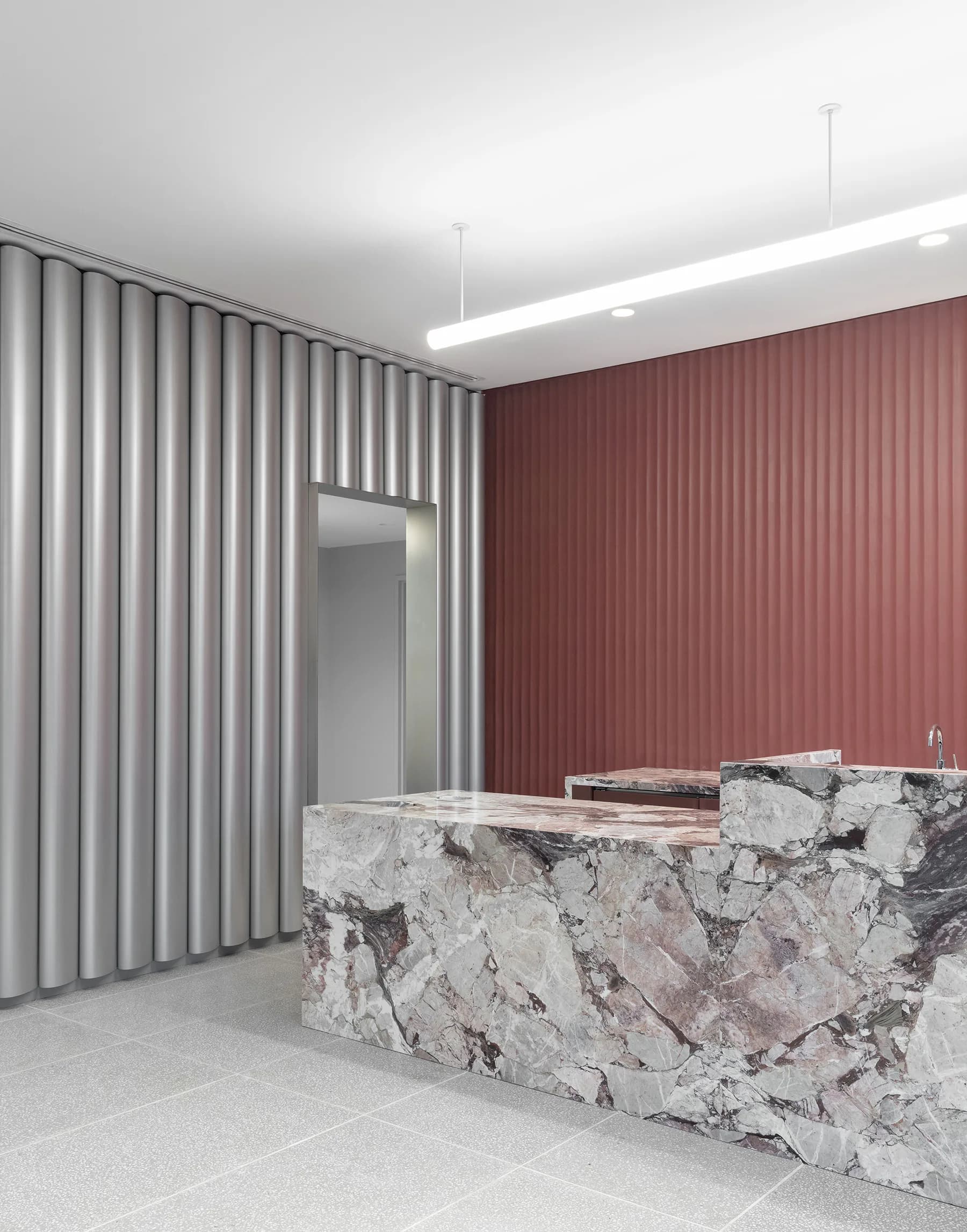 Modern reception space with marble desk, burgundy ribbed wall paneling, vertical striations on left partition, and minimalist ceiling lighting by Dominic Leong.