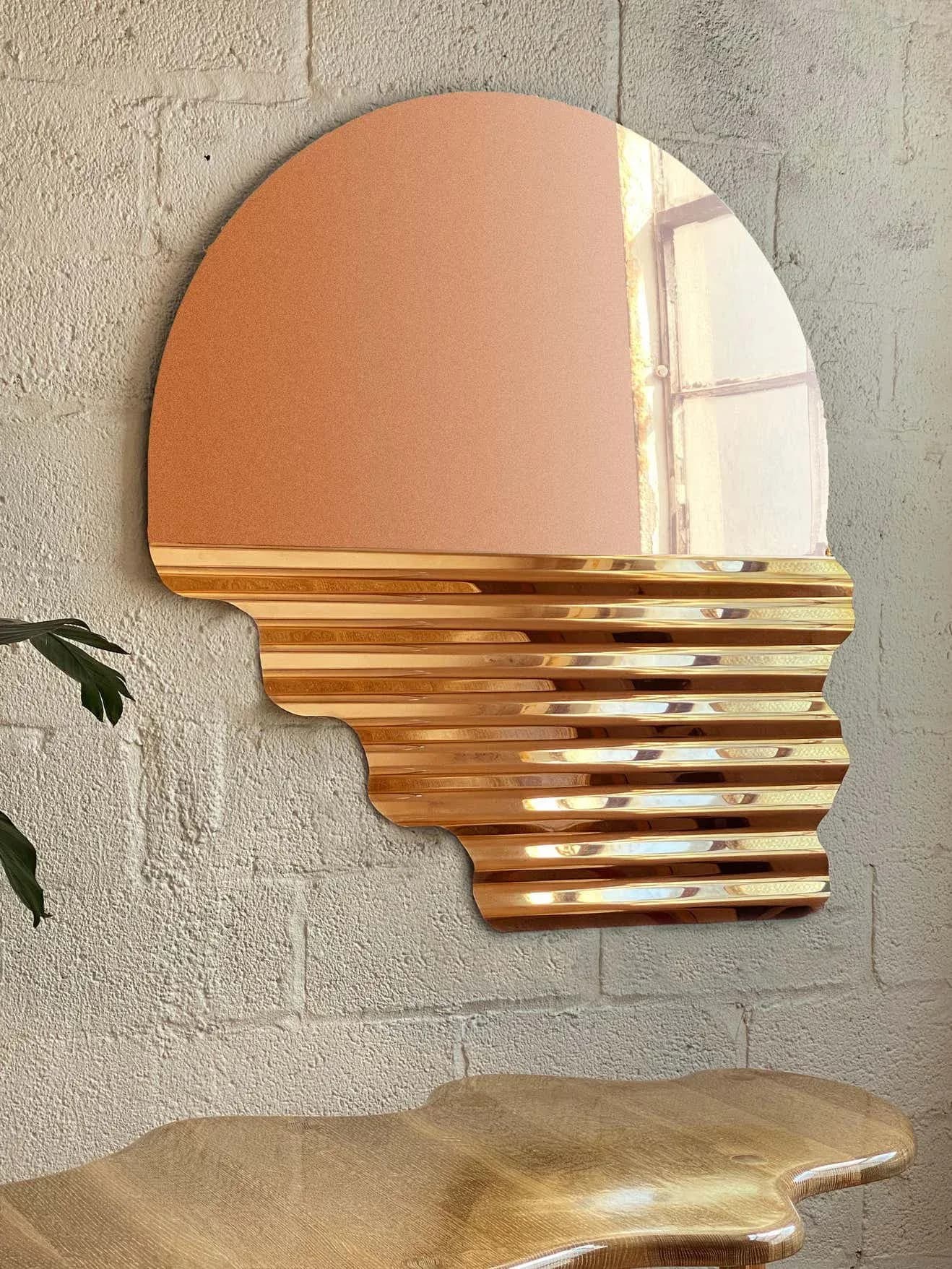 Head-shaped mirror with copper reflective surface and wooden slat lower section mounted on white brick wall, by Dustin John.