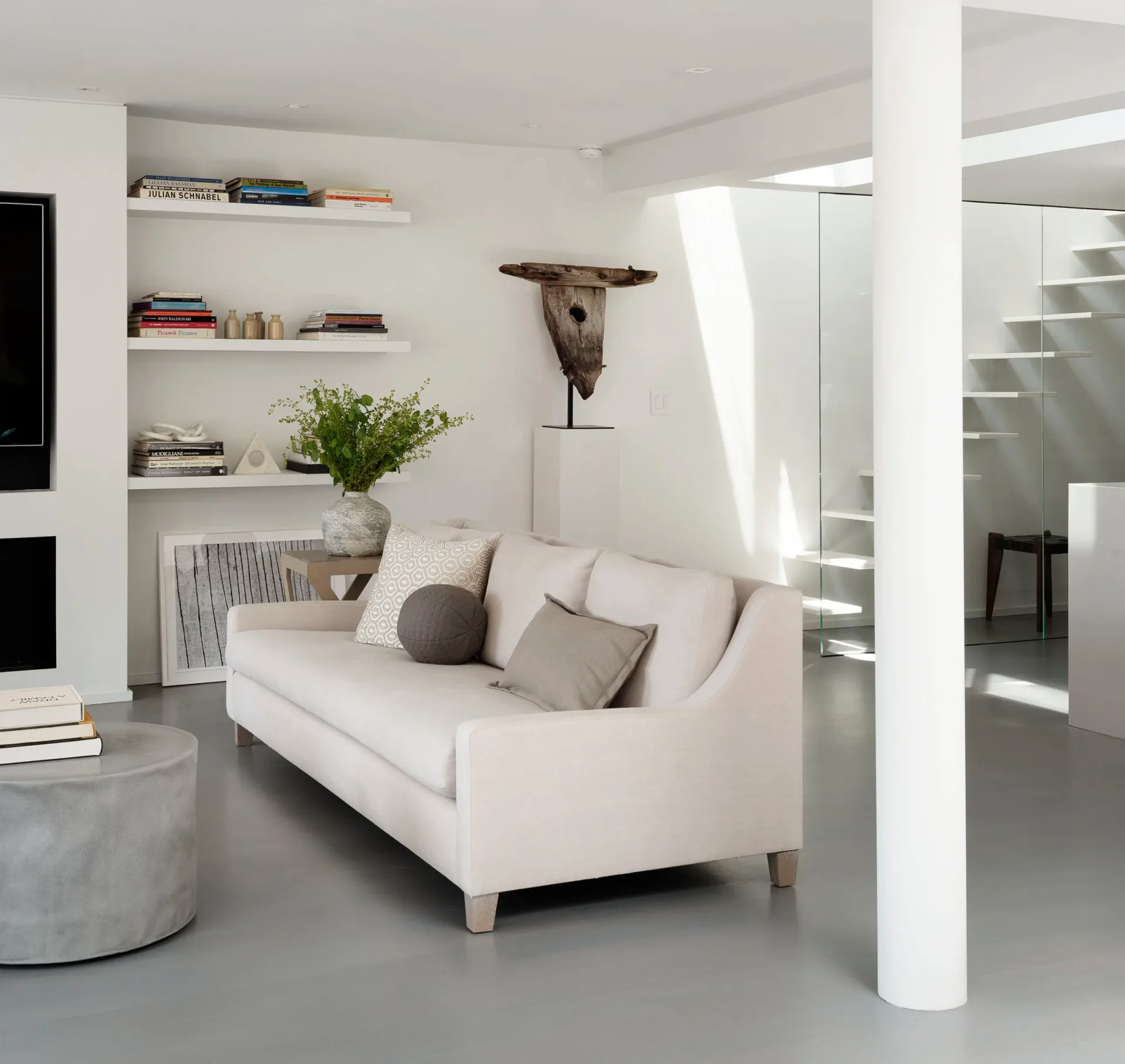 Minimalist living room with cream sofa, floating shelves, potted plants, concrete elements, and bright natural light, designed by Dyana Kazan.