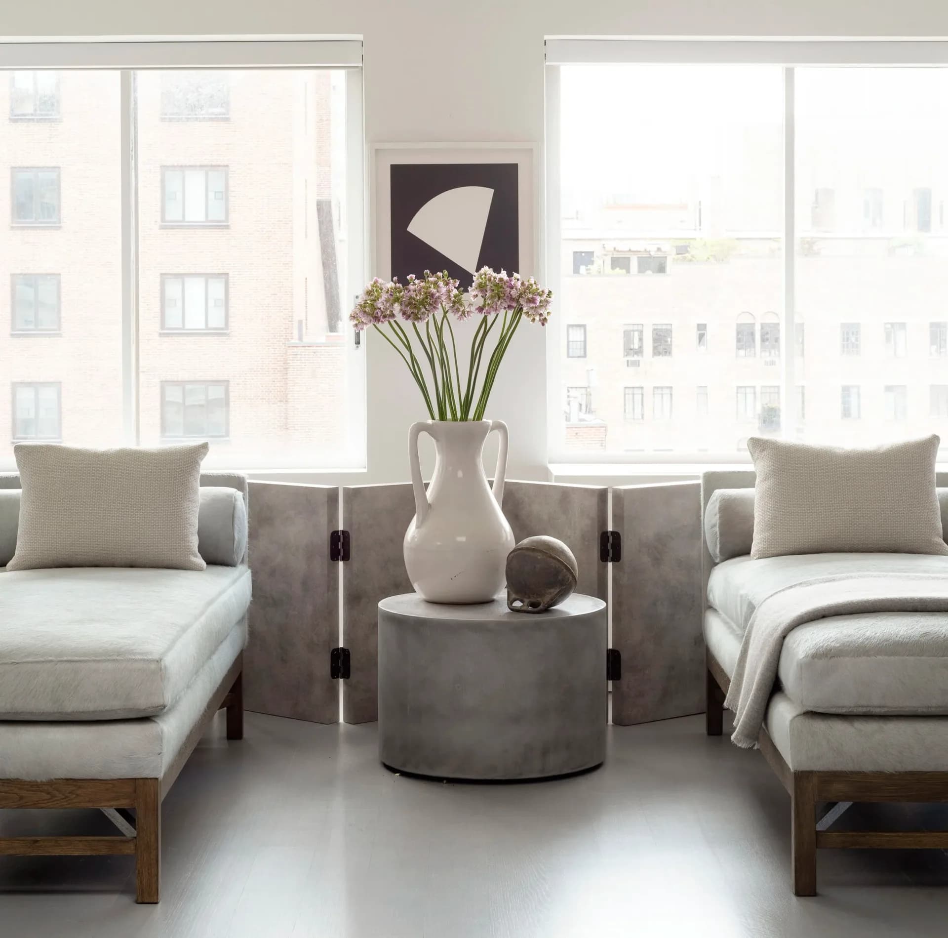 Minimalist living room with symmetrical seating, concrete center table, white vase with flowers, large windows overlooking cityscape by Dyana Kazan.