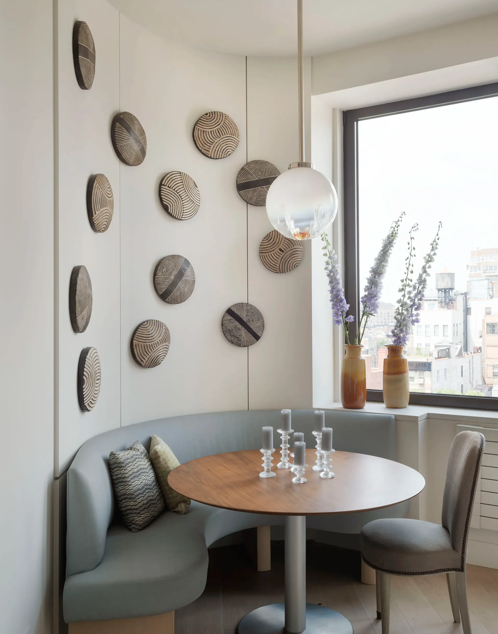 Modern dining nook with curved gray banquette, round wooden table, pendant lighting, and curated wall display of woven circular baskets by Dyana Kazan.