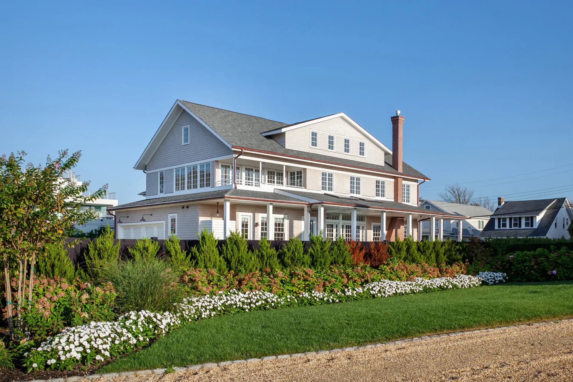 Coastal colonial residence with white trim, wraparound porch, and manicured gardens featuring white flowering plants along the lawn, designed by Ed Kopel.