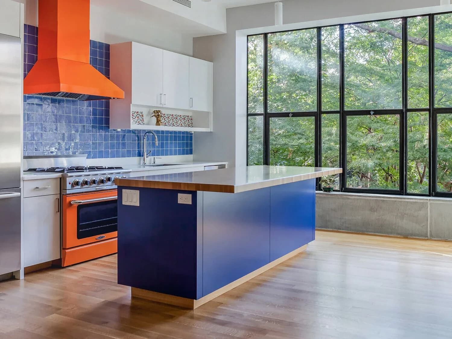 Contemporary kitchen with orange range hood, blue tile backsplash, navy island, and extensive black-framed windows overlooking greenery by Ed Kopel.