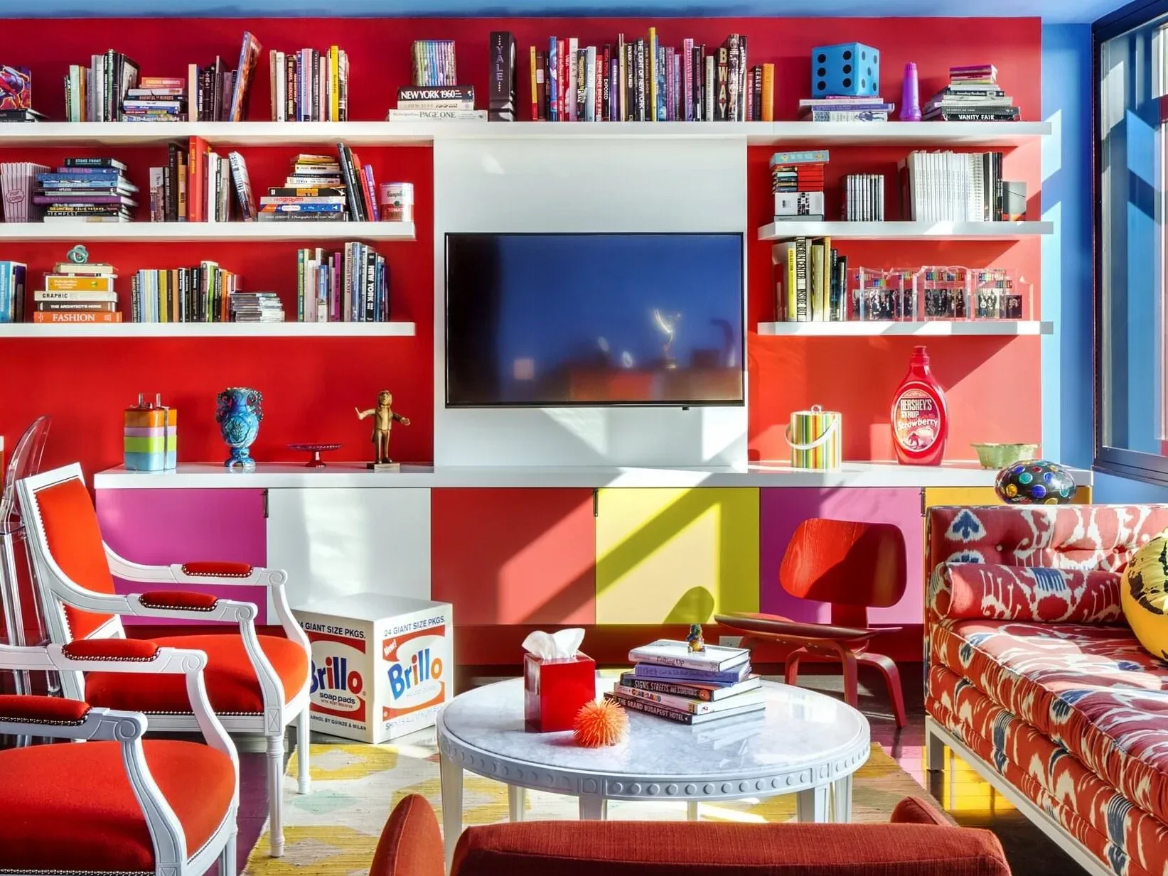 Vibrant red living room with built-in shelving, colorful geometric seating, and media wall by Ed Kopel.