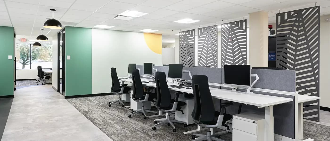 Modern open-plan office space with black task chairs, white desks, green accent walls, geometric patterned panels, pendant lighting, and light wood flooring by Edelyn De La Cruz.