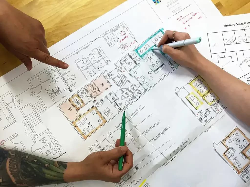 Architectural floor plans with colored annotations and markups being reviewed by hands holding markers during design development.