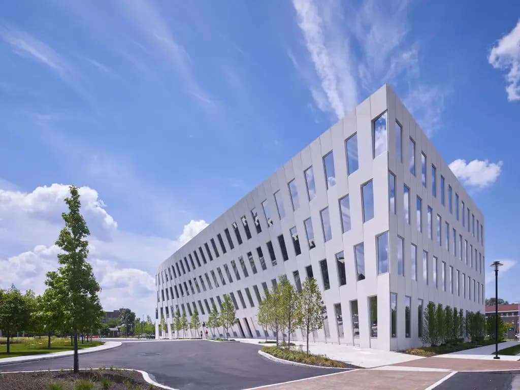 Modern white institutional building with curved corner facade, regular window grid, and landscaped grounds with trees under bright blue sky by Edelyn De La Cruz.