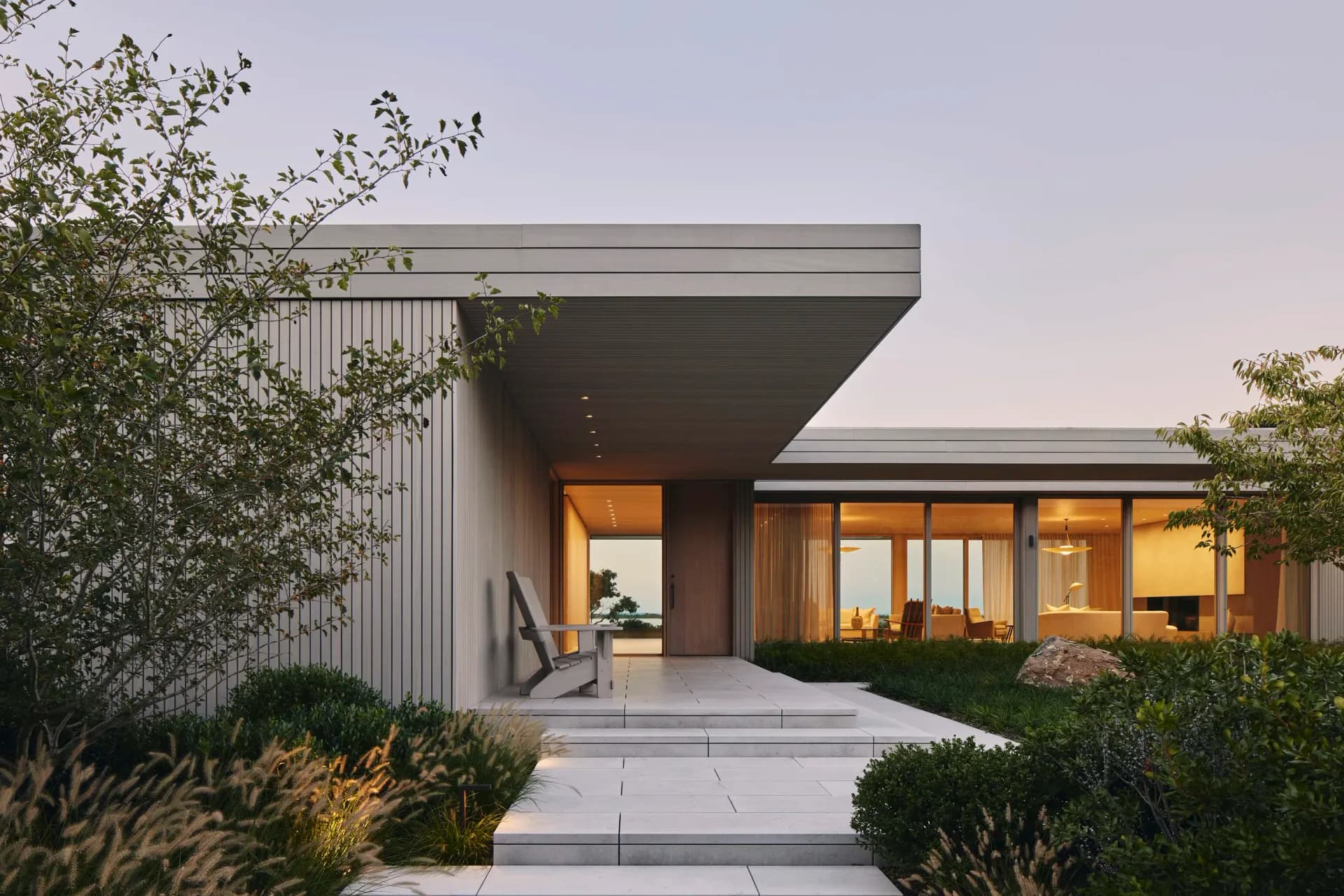 Modern residential exterior with cantilevered roofline, warm interior lighting, horizontal metal cladding, and minimalist landscaping by Eleanor Donnelly.