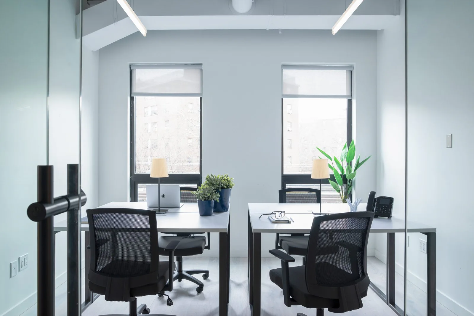 Modern office space with two workstations featuring black mesh chairs, white desks, and large windows with natural light, designed by Elide Rathborne.